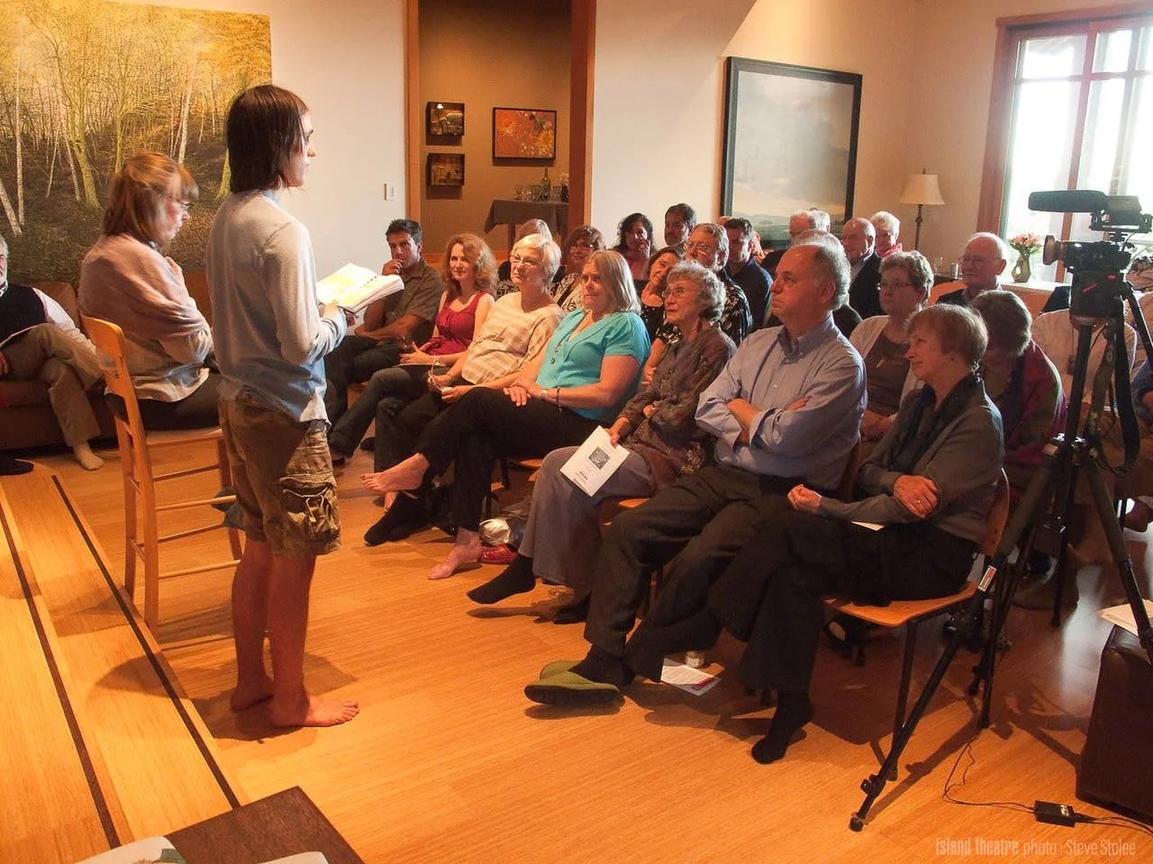 A young person with long dark hair, wearing a white long-sleeve shirt and cargo shorts, stands barefoot in front of an audience, reading from a paper. The audience, consisting mostly of elderly adults, is seated and attentively listening in a well-lit room with artwork on the walls and a large window.