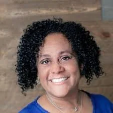 A woman with curly black hair, smiling, wearing a blue top, in front of a wooden background.