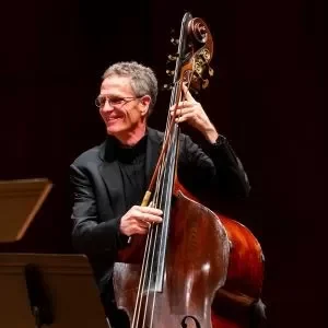 A man smiling and playing a double bass on stage, wearing a black suit.