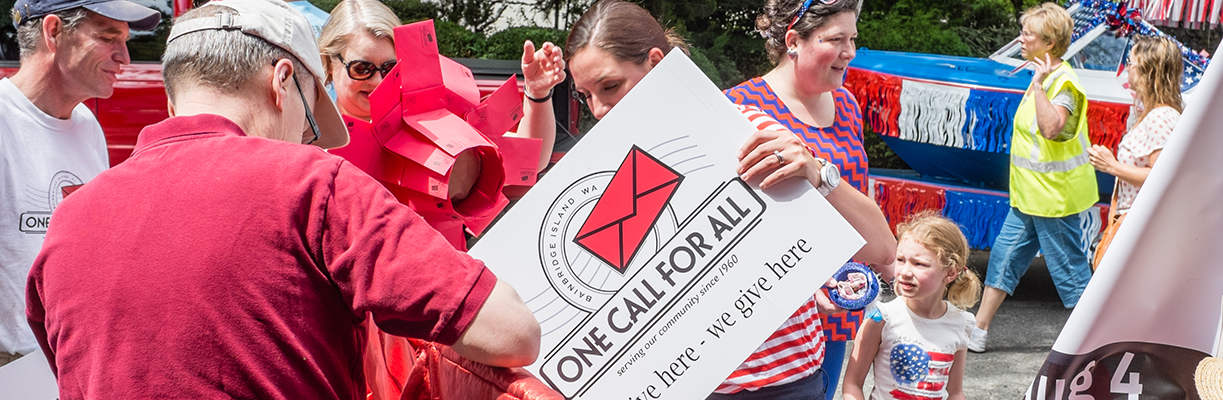People participating in an outdoor event, involving a donation drive with a large check and red donation boxes, with a float decorated in red, white, and blue in the background.