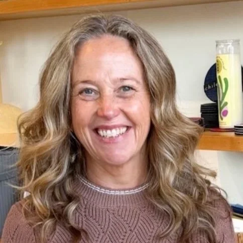 A smiling woman with long, curly blonde hair wearing a brown sweater in an indoor setting, with a shelf and various items in the background.