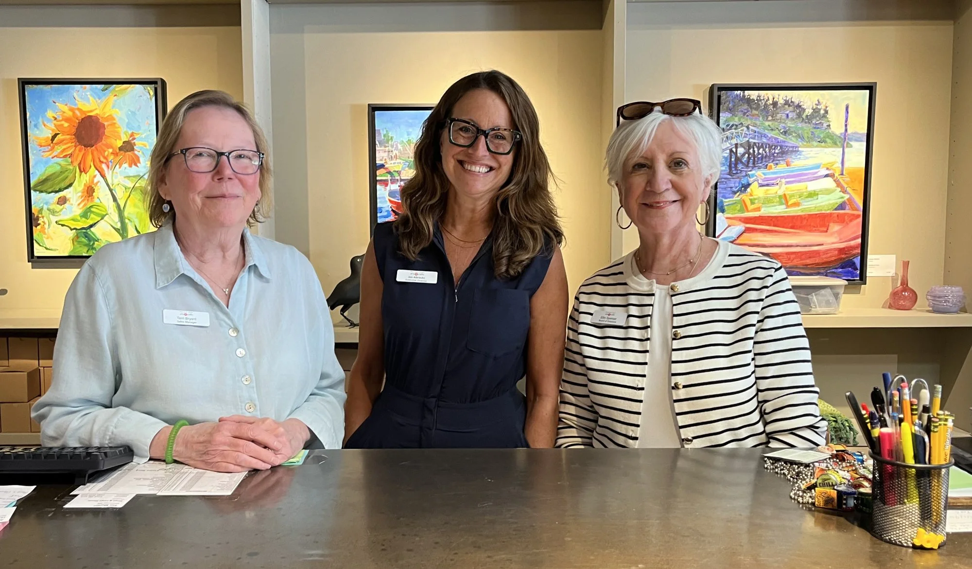 Three women standing behind a counter in an art gallery or studio, with colorful paintings in the background. They are smiling and wearing name tags.