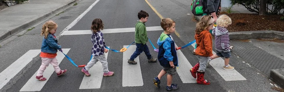 Six children holding hands and crossing the street on a pedestrian crosswalk, with an adult supervising in the background.
