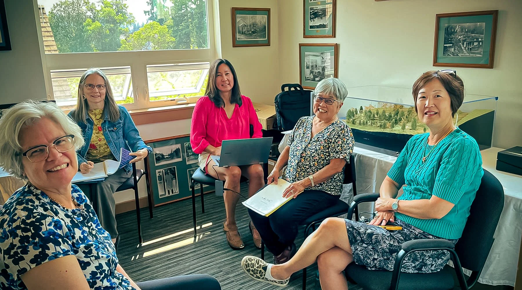 Group of five women sitting in a conference room with windows and various framed pictures on the walls, engaged in a meeting or discussion.