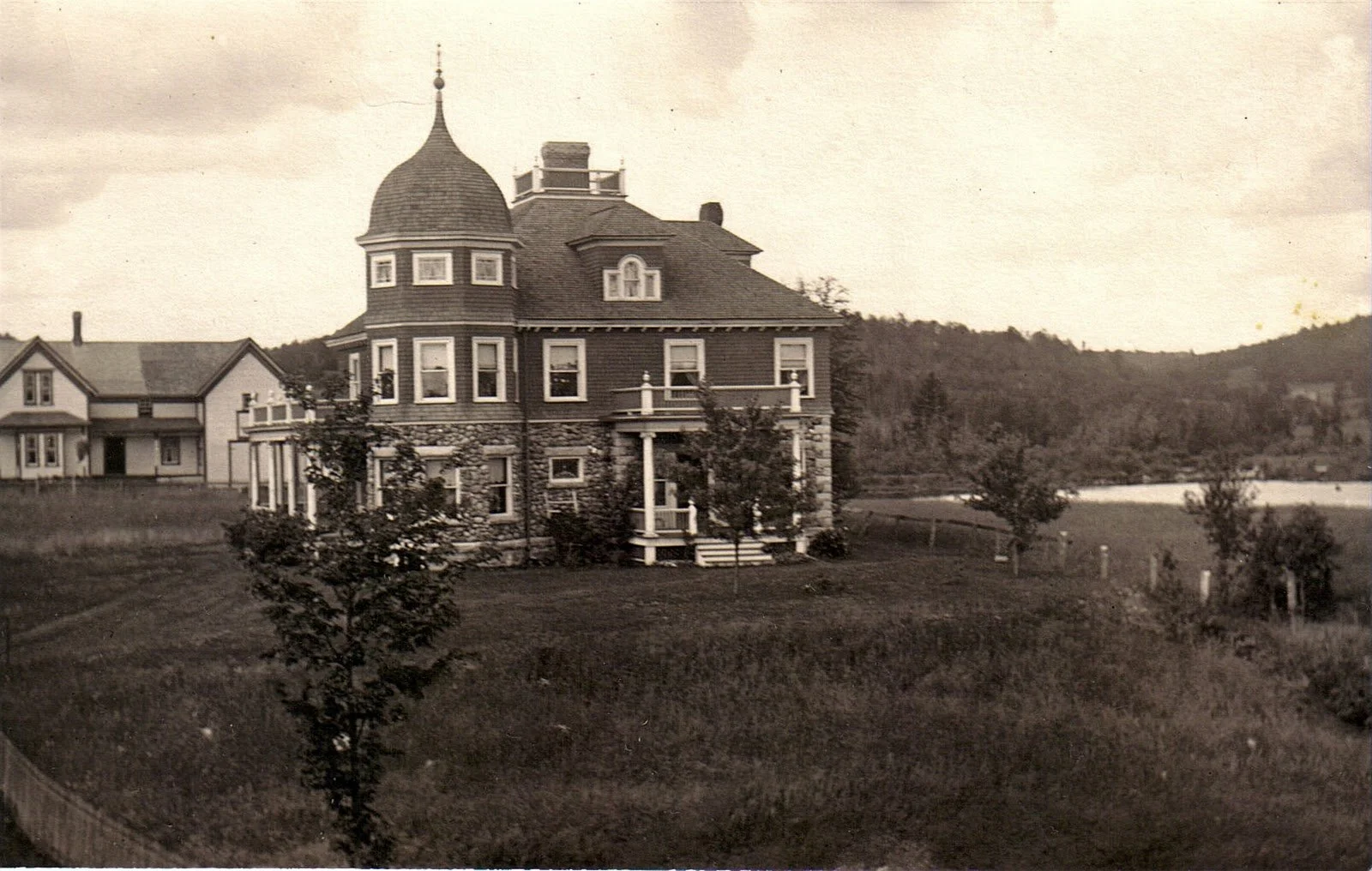 An old black and white photograph of a large Victorian-style house with a turret, set on a grassy lawn with small trees and a hill with trees in the background.