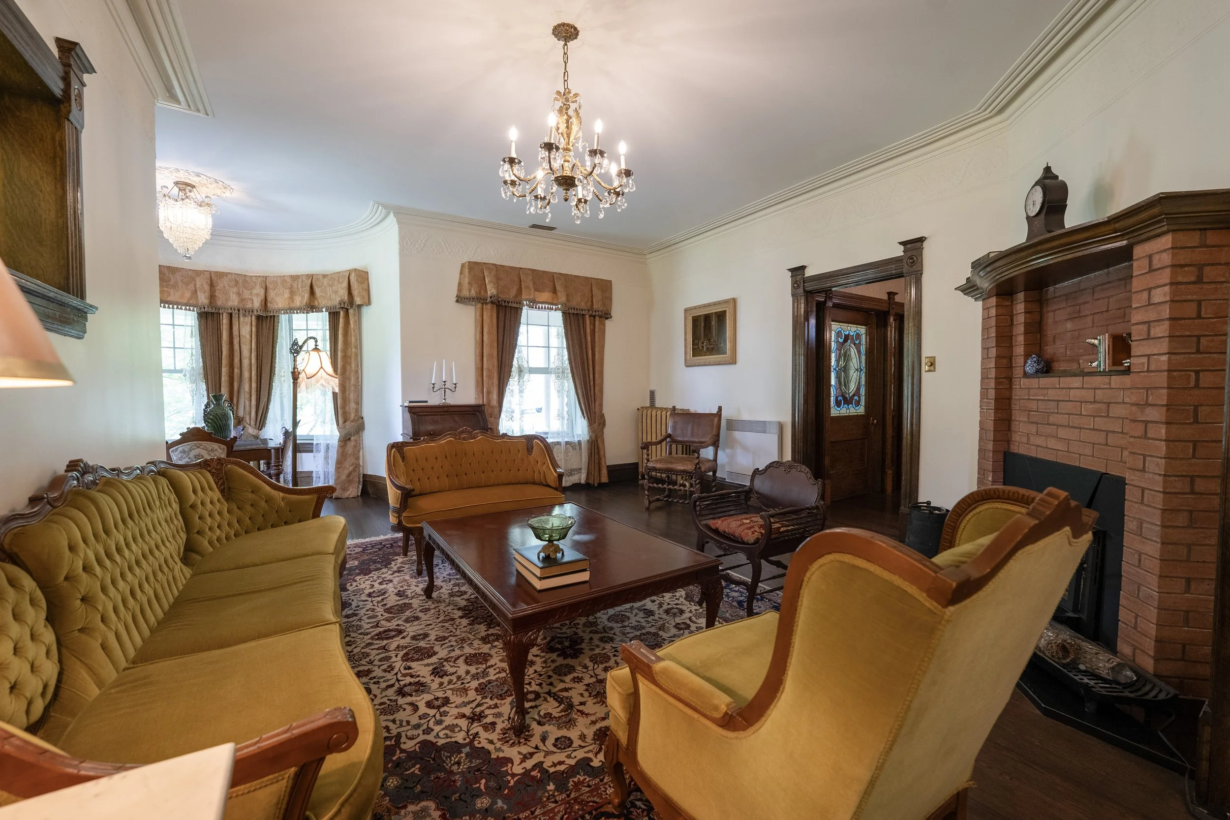 Living room with vintage yellow sofas, wooden coffee table, area rug, large windows with curtains, chandelier, a brick fireplace, and antique decor.