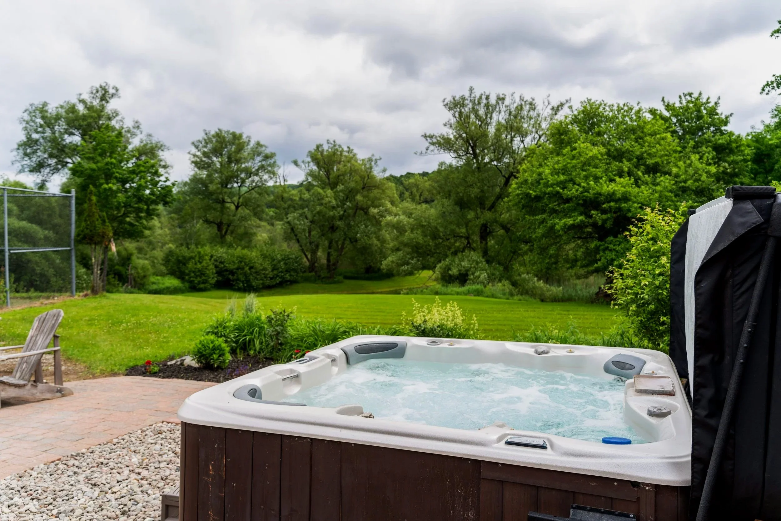 Hot tub on a patio with a lush green backyard and cloudy sky.