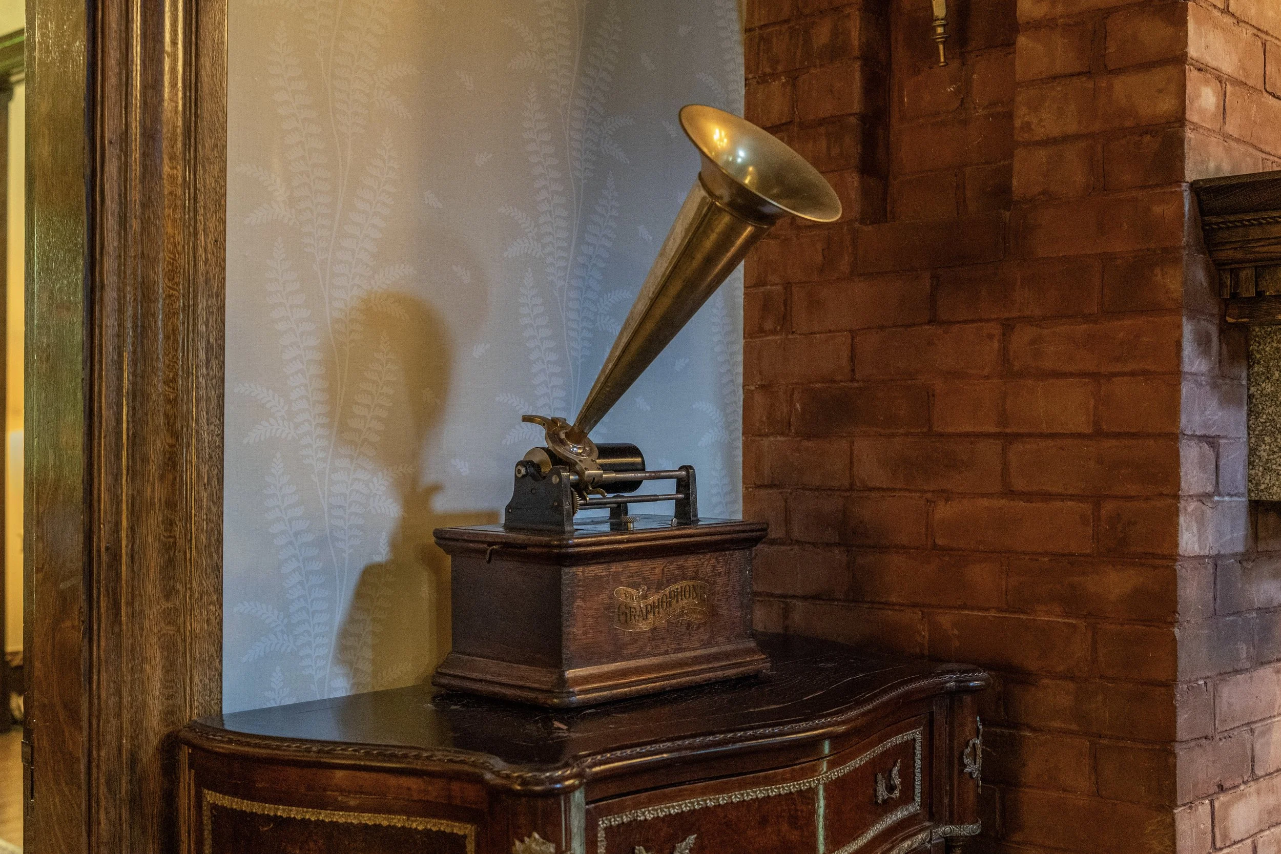 An antique phonograph with a large brass horn on a wooden cabinet, positioned on a dark wooden table against a brick wall and wallpapered wall with fern patterns.