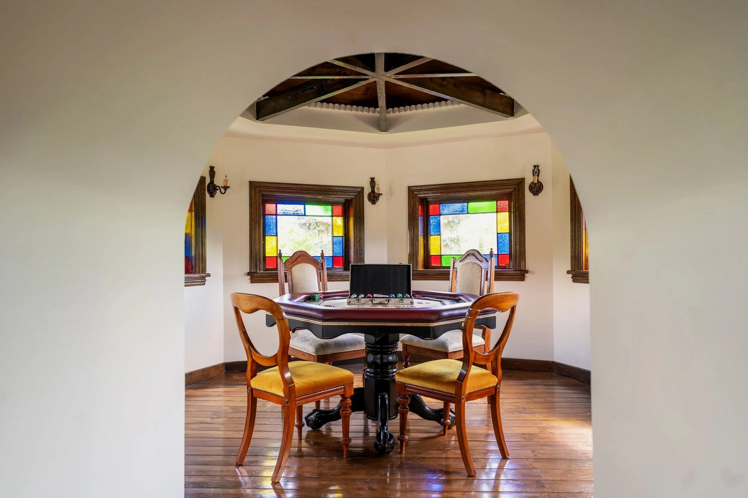 A dining room with a round wooden table surrounded by four wooden chairs with yellow upholstered seats, two stained glass windows with red, blue, yellow, and green panels, and a wooden ceiling with exposed beams.