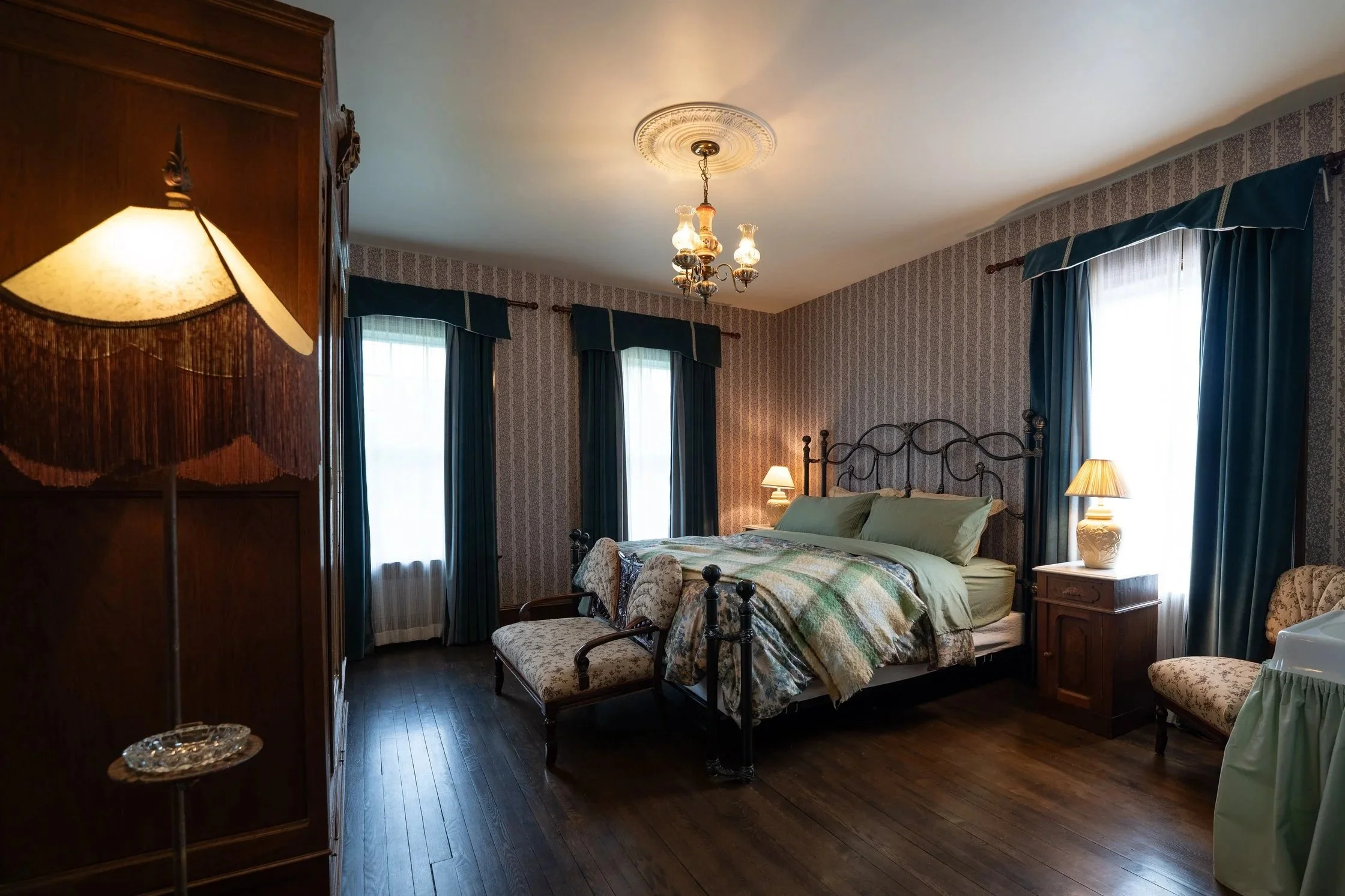 A vintage-style bedroom with a four-poster bed, bedside lamps, dark wood furniture, blue curtains, patterned wallpaper, and a chandelier hanging from the ceiling.