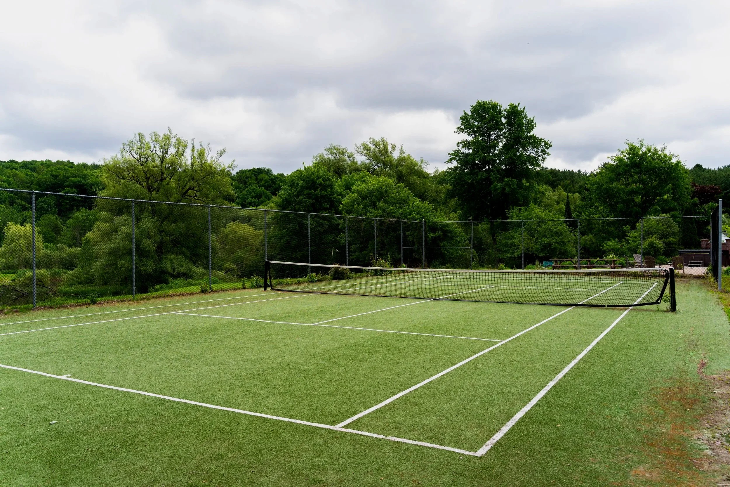 An outdoor tennis court with a green surface and white lines, surrounded by a chain-link fence, with trees and cloudy sky in the background.
