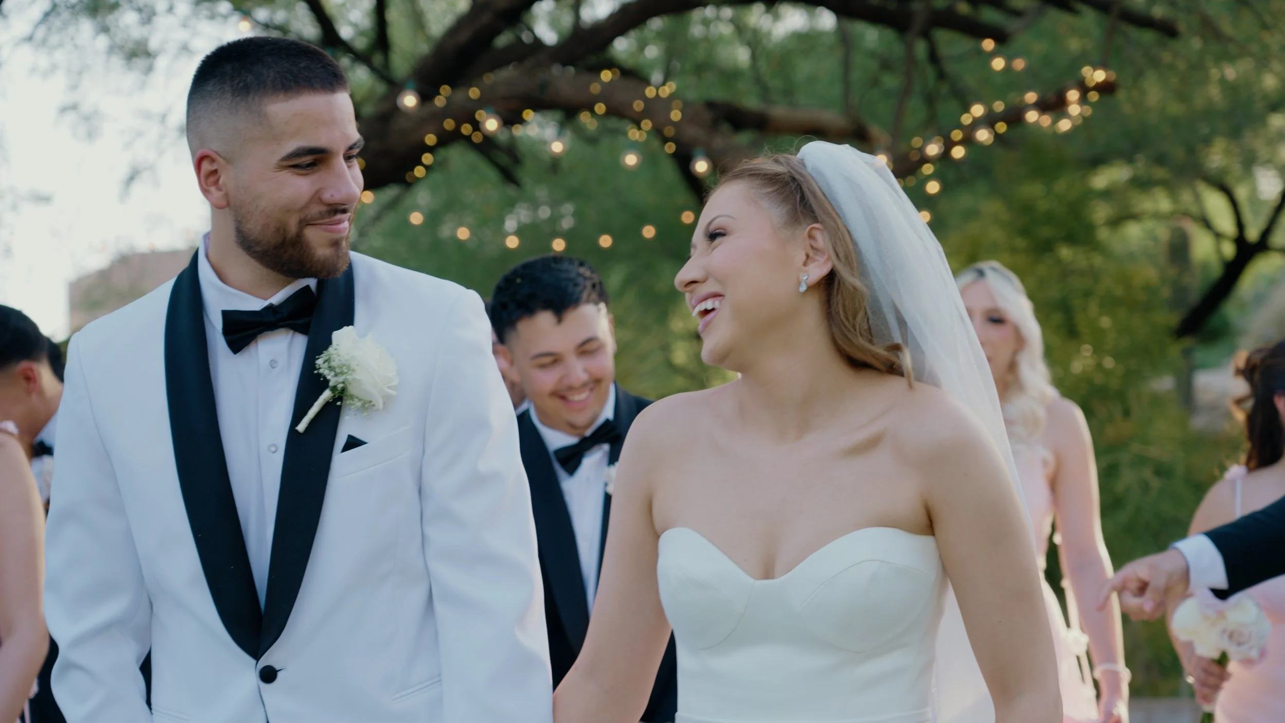 A bride and groom smile at each other during their wedding celebration outdoors, with string lights hanging among trees in the background.