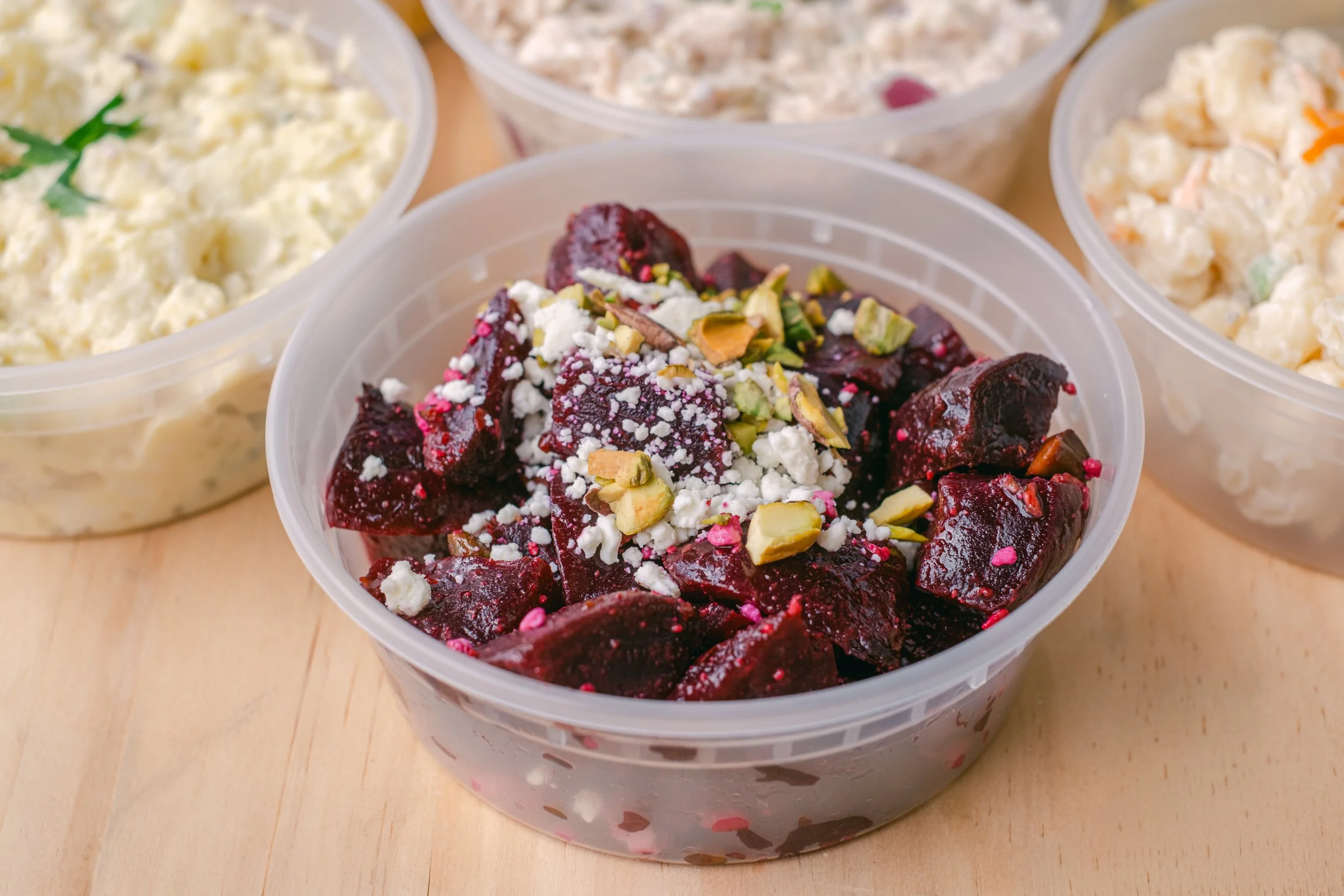 Plastic containers with potato salad, chicken salad, and beet salad on a wooden surface.
