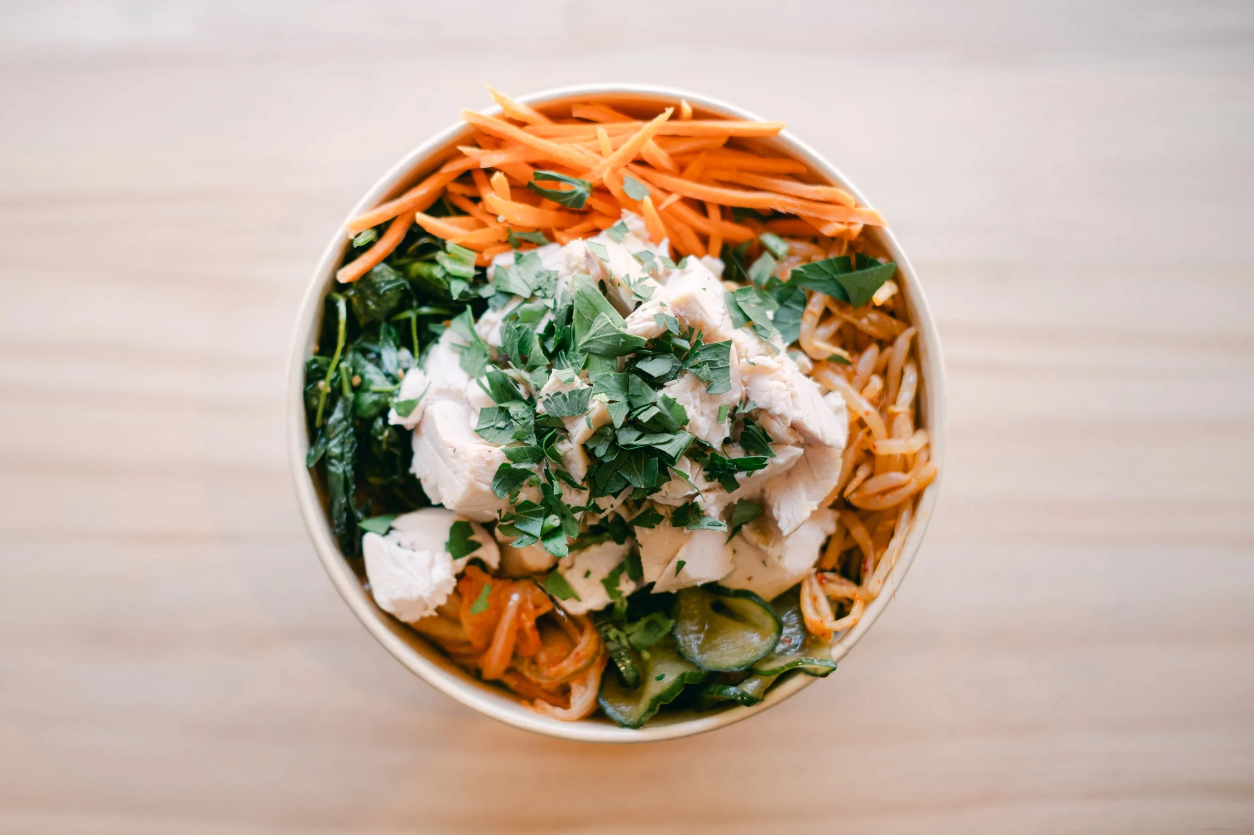 A bowl of Asian noodle salad with shredded carrots, cooked chicken, chopped greens, sliced cucumbers, and fresh herbs.