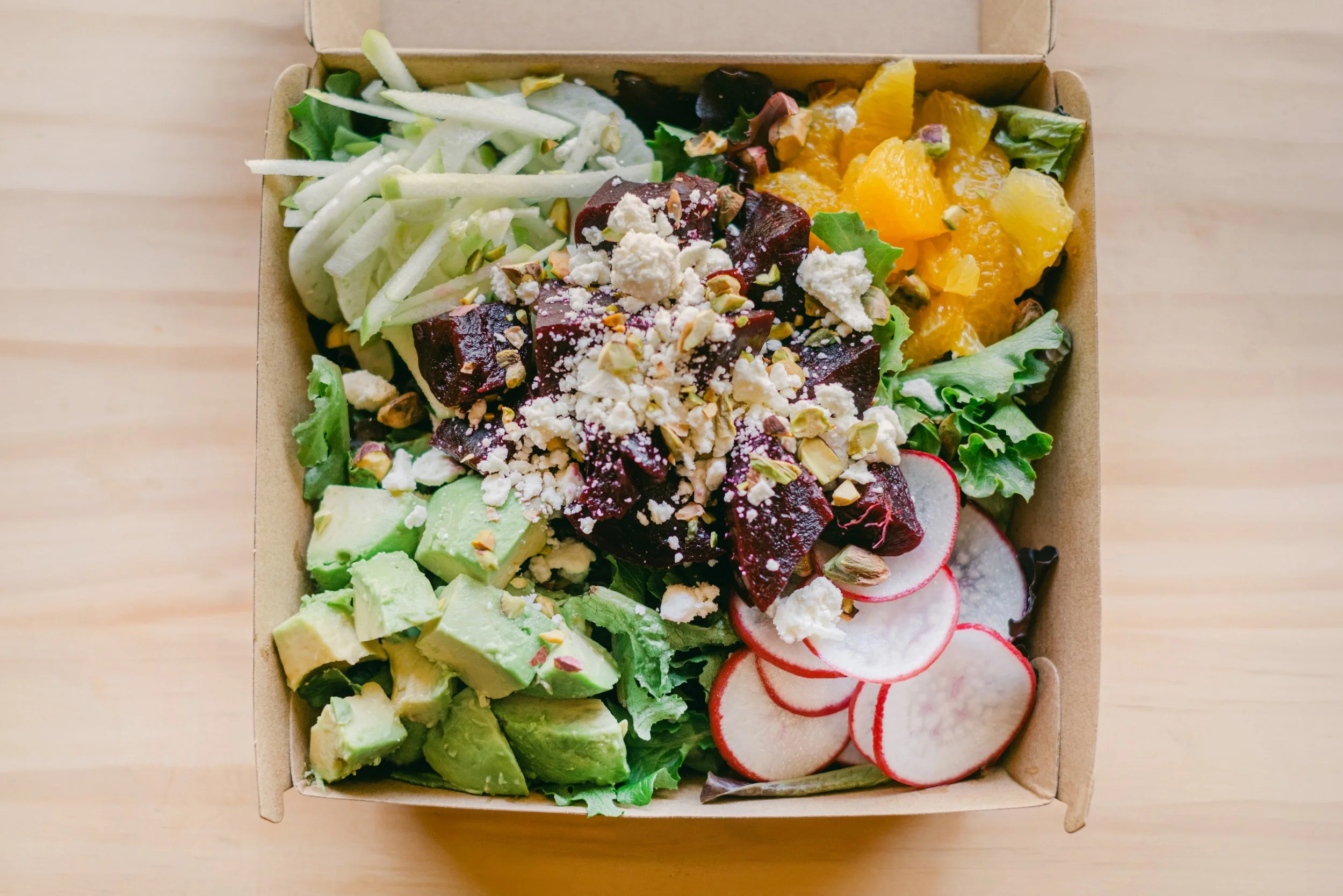 A cardboard box filled with fresh salad ingredients including sliced cucumbers, radishes, avocado, chopped greens, orange and yellow citrus segments, beets, crumbled white cheese, and chopped nuts.