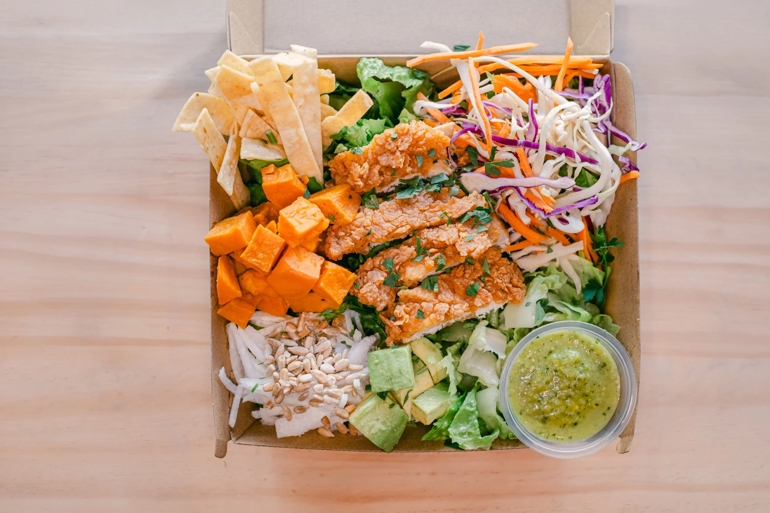 Takeout box with assorted salad ingredients including grilled chicken, shredded cabbage, carrots, purple cabbage, lettuce, avocado, sweet potatoes, sunflower seeds, shredded cheese, tortilla strips, a small container of green dressing, and sunflower 