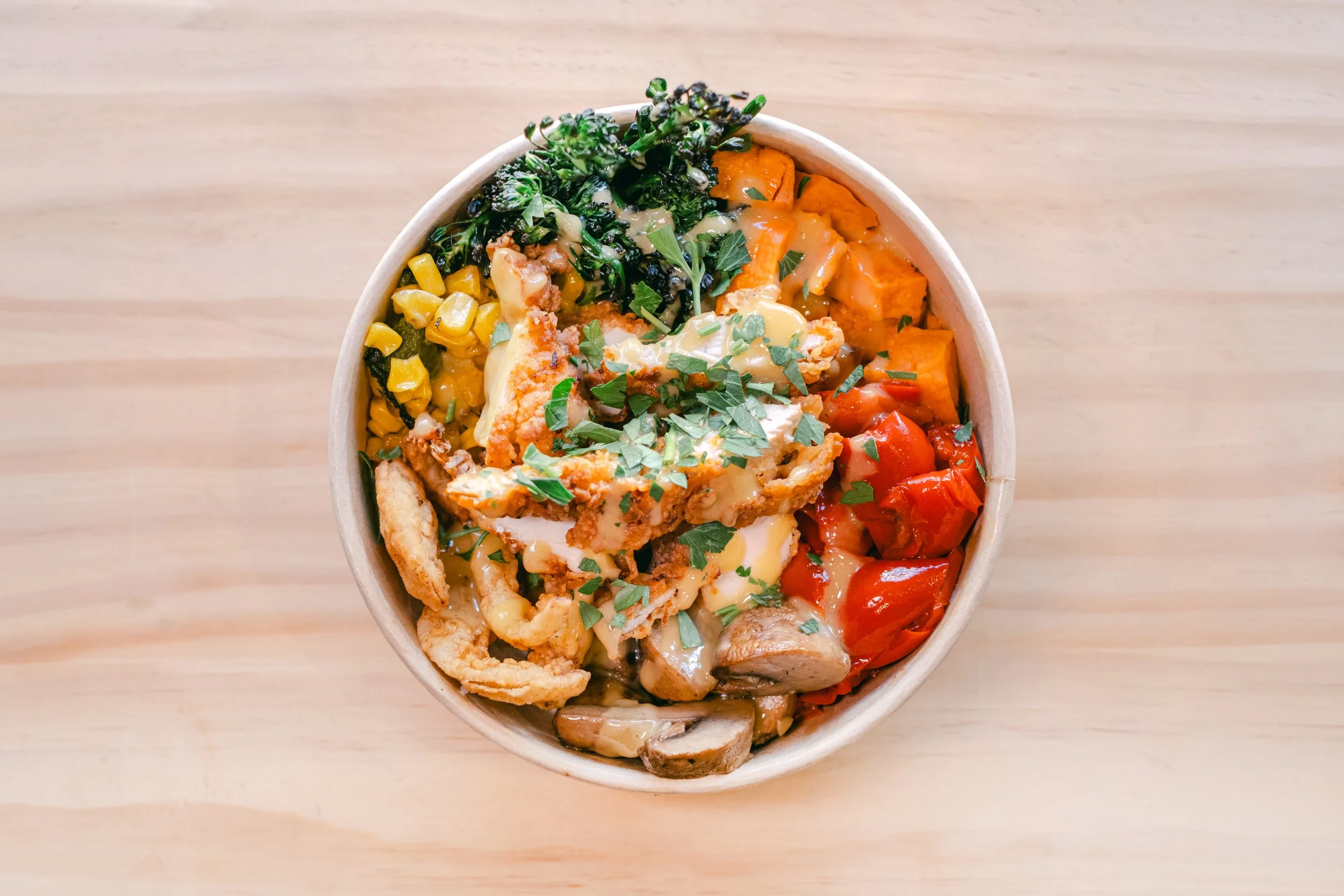 A bowl of food filled with vegetables including corn, kale, sweet potatoes, red bell peppers, and mushrooms, garnished with chopped herbs.