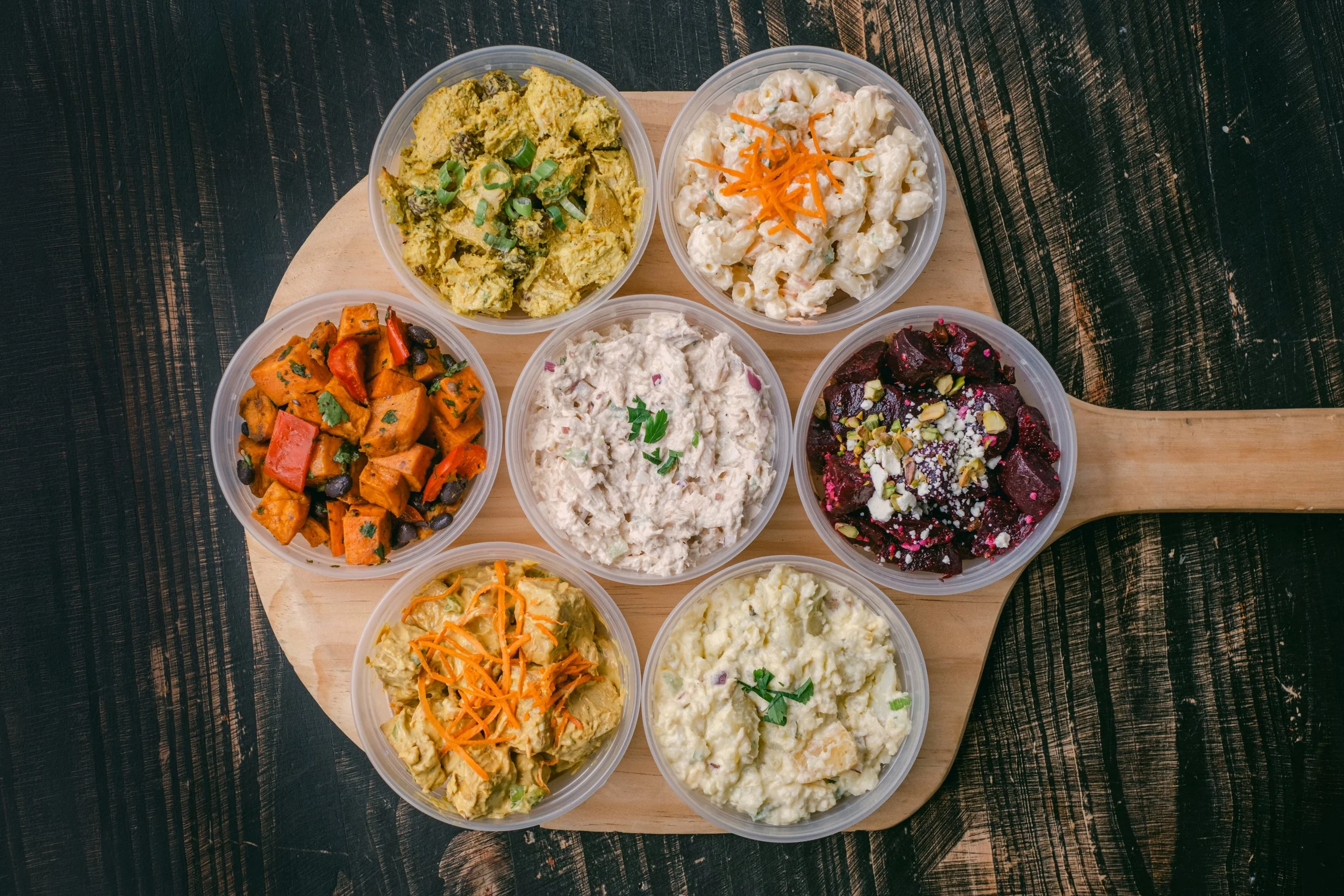 Assortment of nine different potato salads and side dishes in clear plastic containers on a wooden serving board.