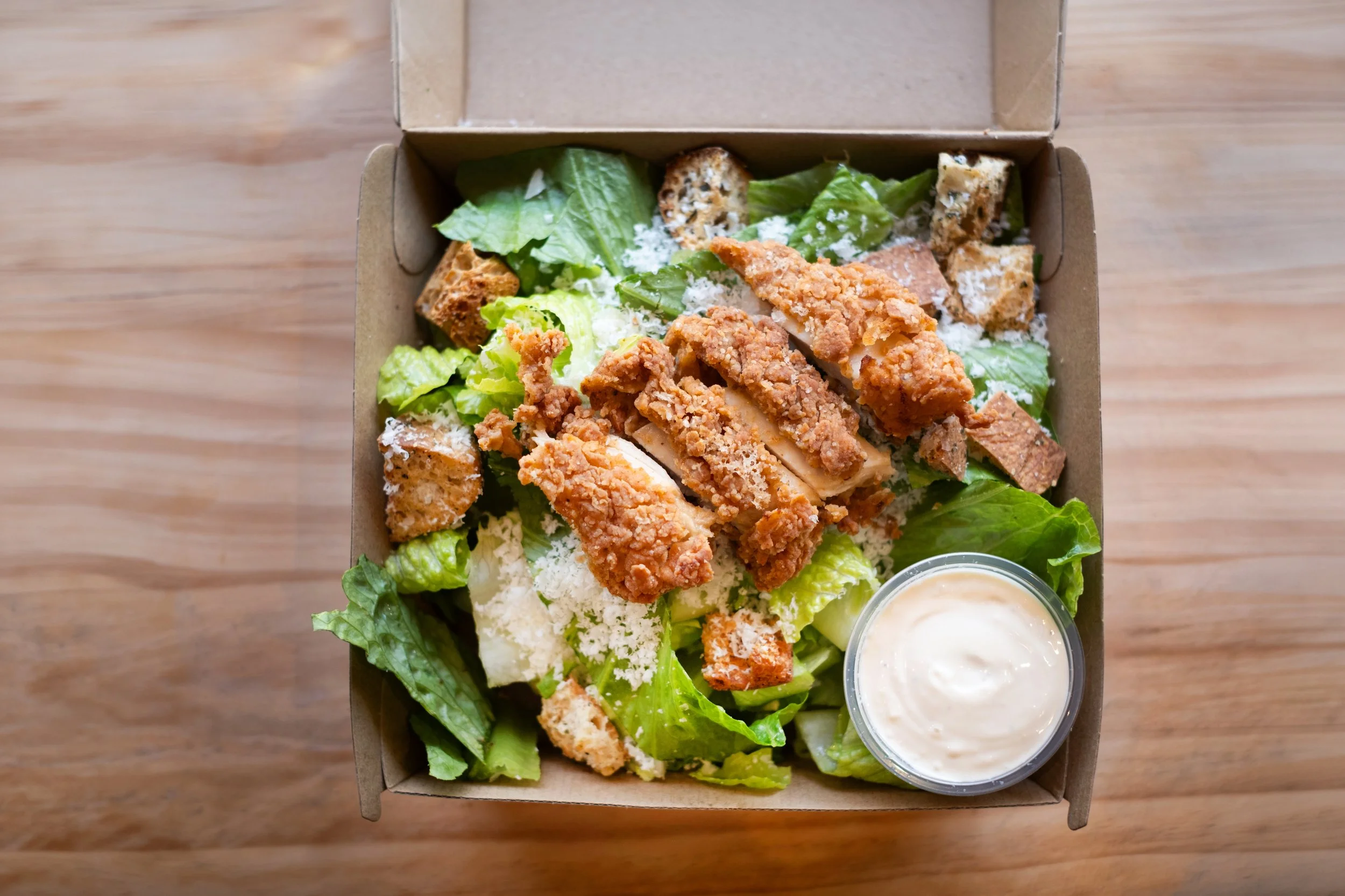 A takeout cardboard box filled with a salad containing crispy fried chicken strips, romaine lettuce, shredded cheese, croutons, and two small cups of dressing.