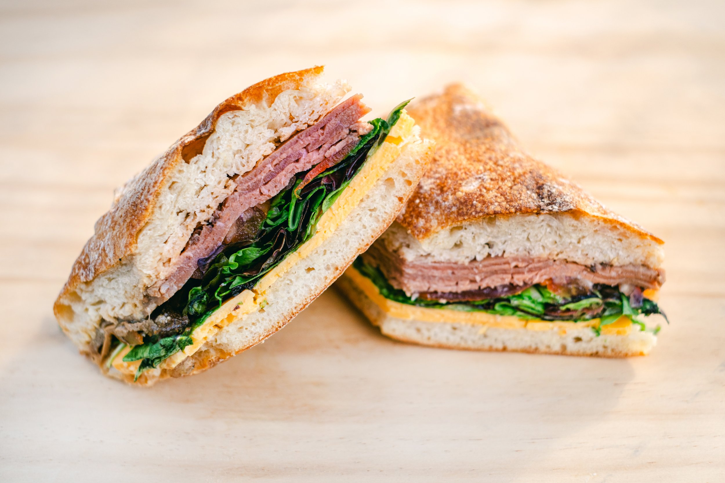 Cross-section of a sandwich with bread, sliced deli meat, lettuce, tomato, and cheese on a wooden surface.