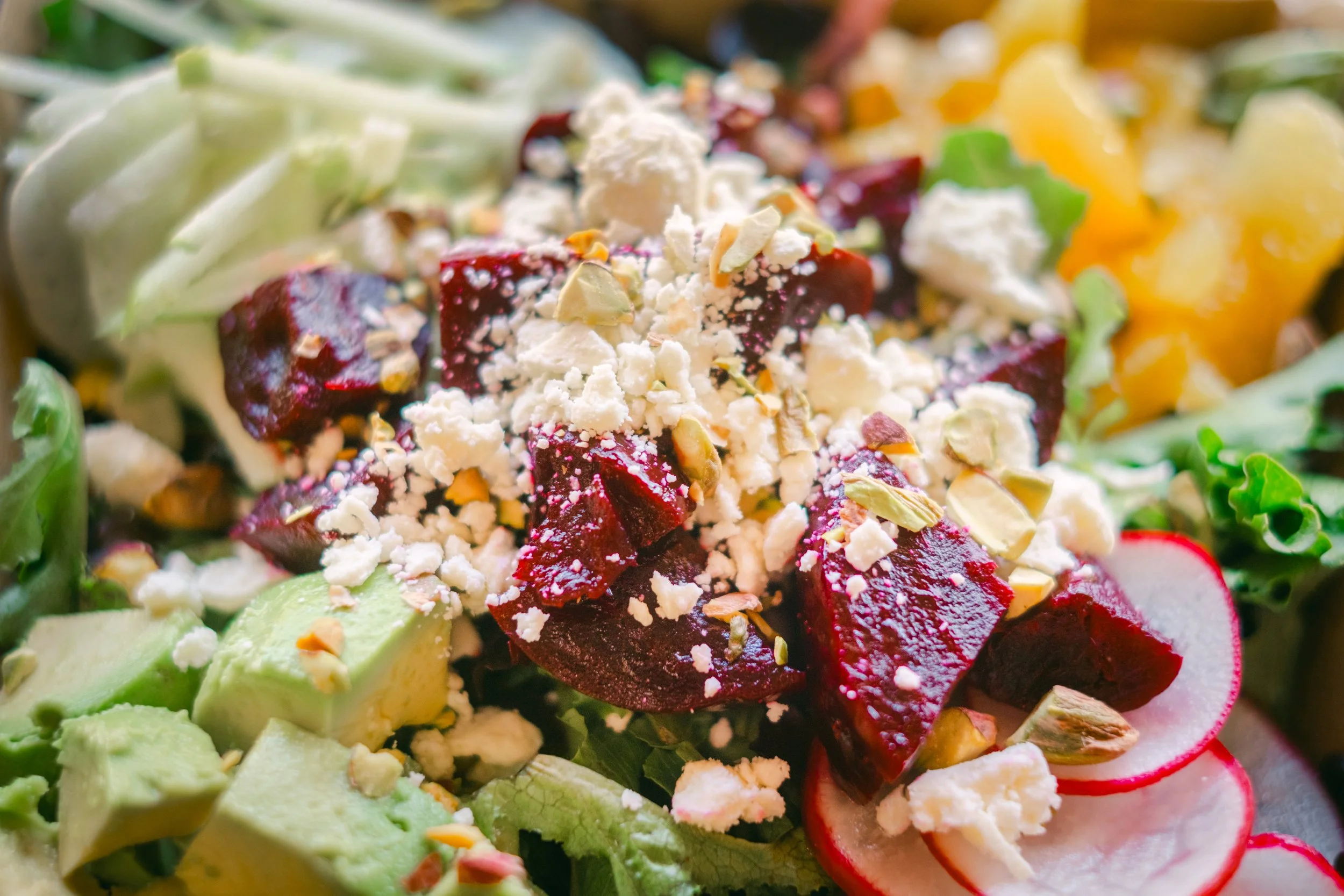 close-up of a fresh vegetable salad with slices of radish, avocado, and diced beets topped with crumbled cheese and chopped nuts.