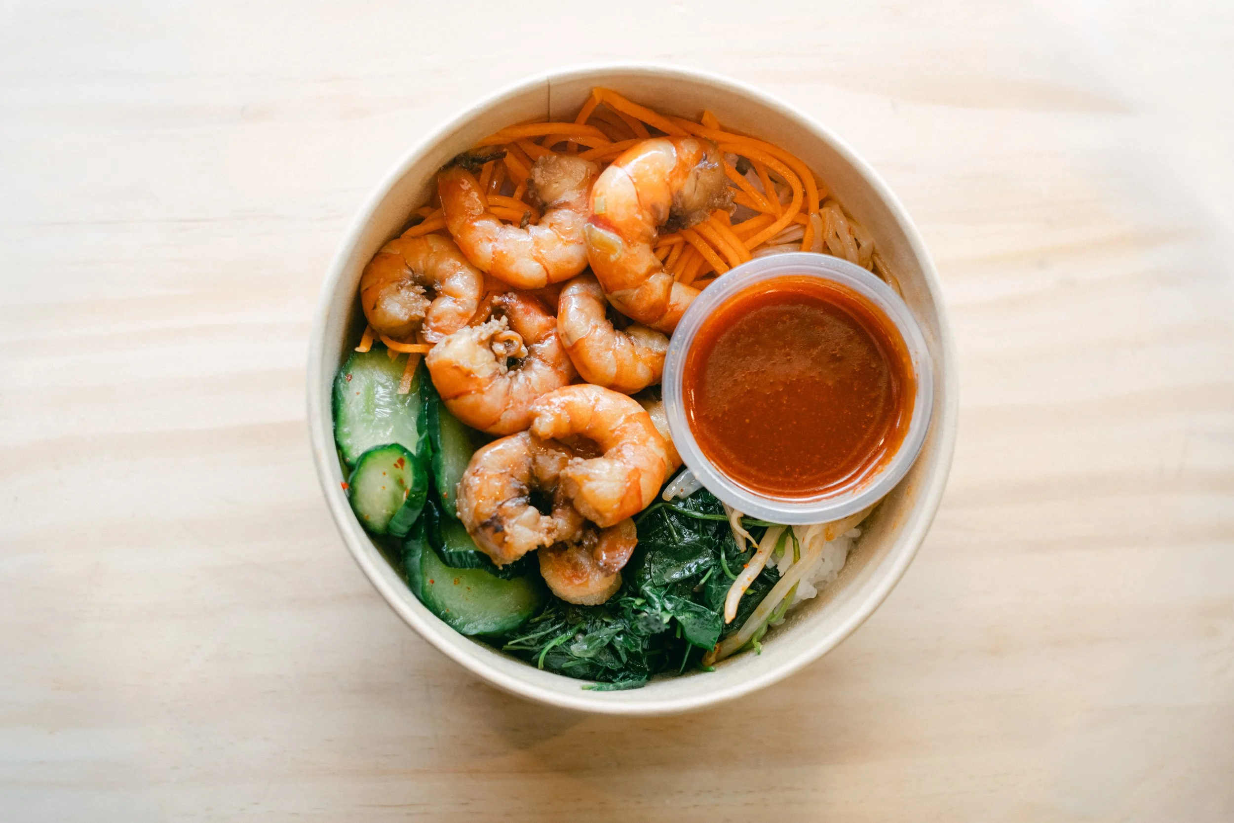 A takeout bowl containing shrimp, noodles, vegetables, and a small container of brown sauce, placed on a light-colored wooden surface.
