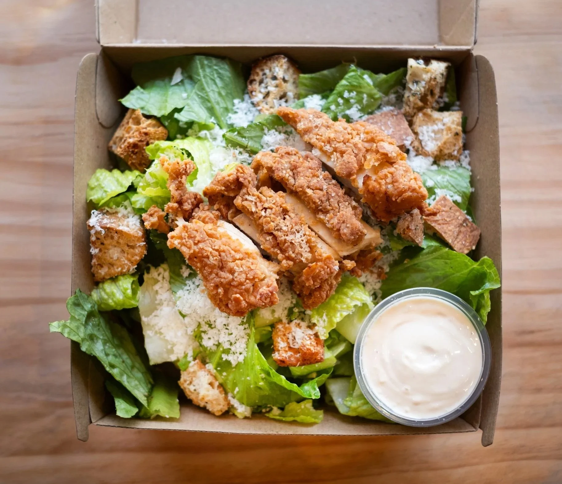 A cardboard container of fried chicken strips, chopped Romaine lettuce, croutons, shredded cheese, and a side of creamy dressing in a small cup.