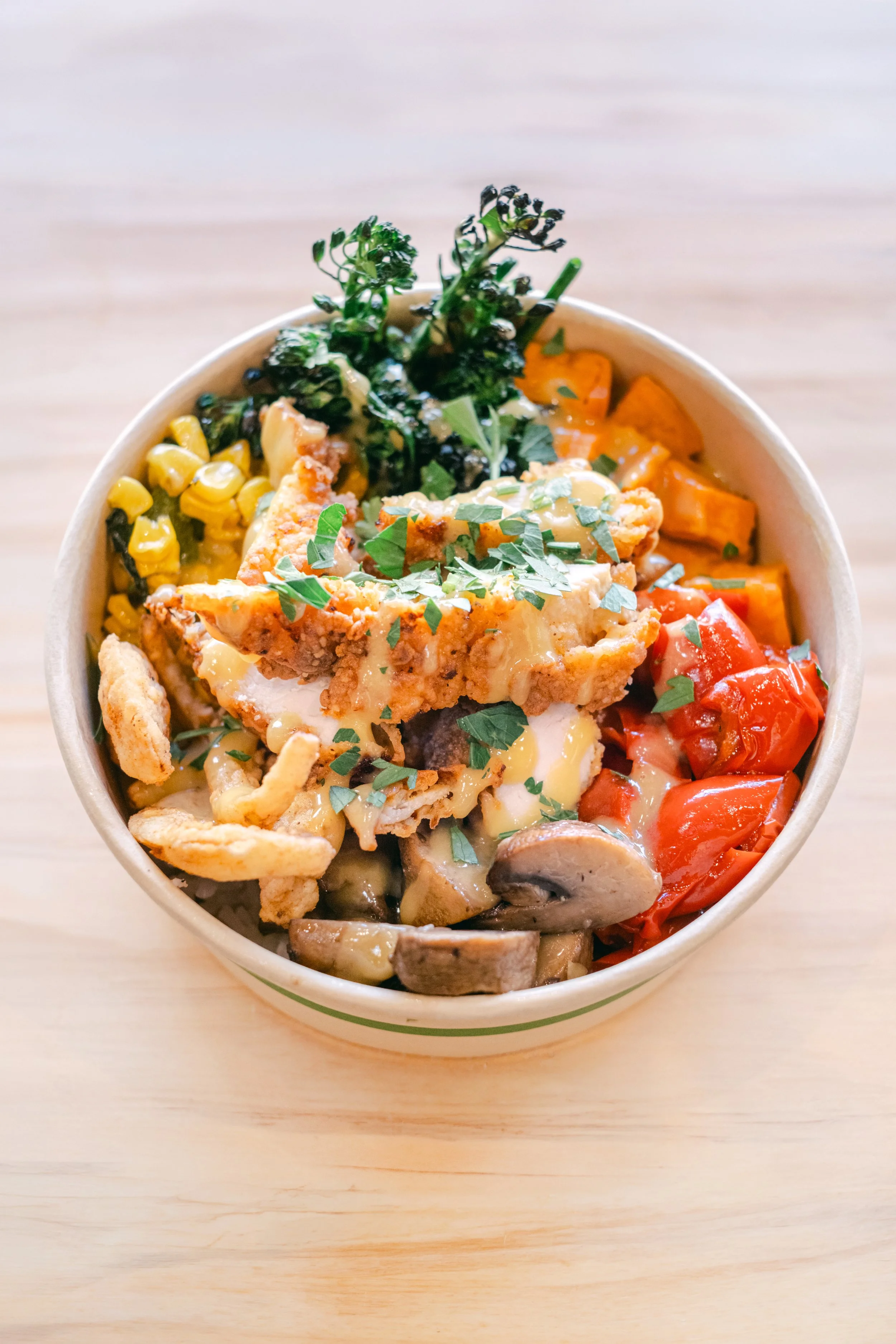 A bowl of rice topped with fried chicken, chopped herbs, and melted cheese, surrounded by roasted vegetables including cherry tomatoes, mushrooms, corn, and sweet potatoes.