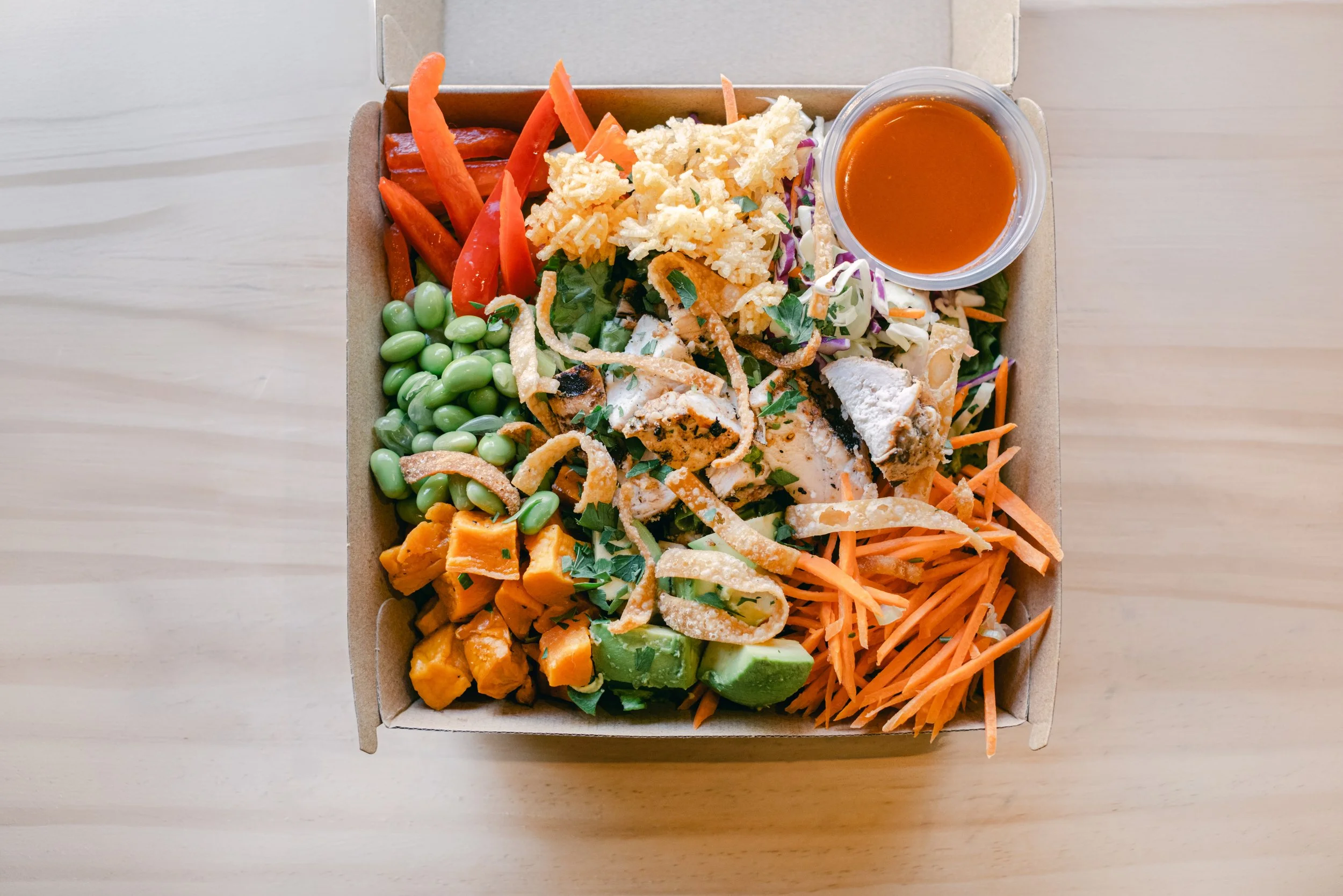 A rectangular cardboard takeout box filled with a colorful salad containing assorted vegetables, shredded cheese, grilled chicken pieces, crispy fried onions, and a side cup of orange salad dressing on a light wooden surface.