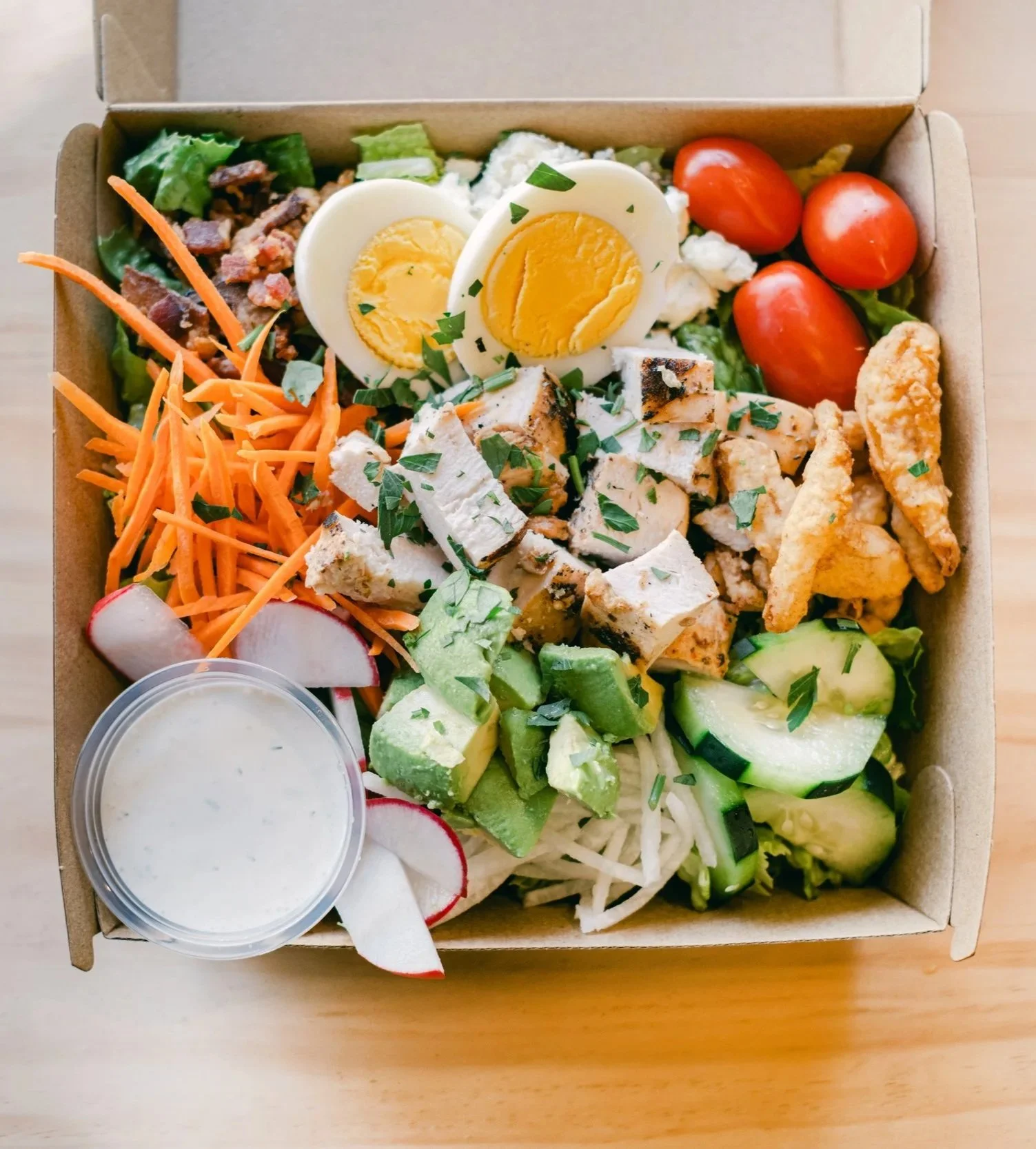 A takeout salad box with cherry tomatoes, sliced radishes, shredded carrots, chopped chicken, sliced cucumbers, grape tomatoes, hard-boiled eggs, crumbled cheese, lettuce, and a side of dressing.