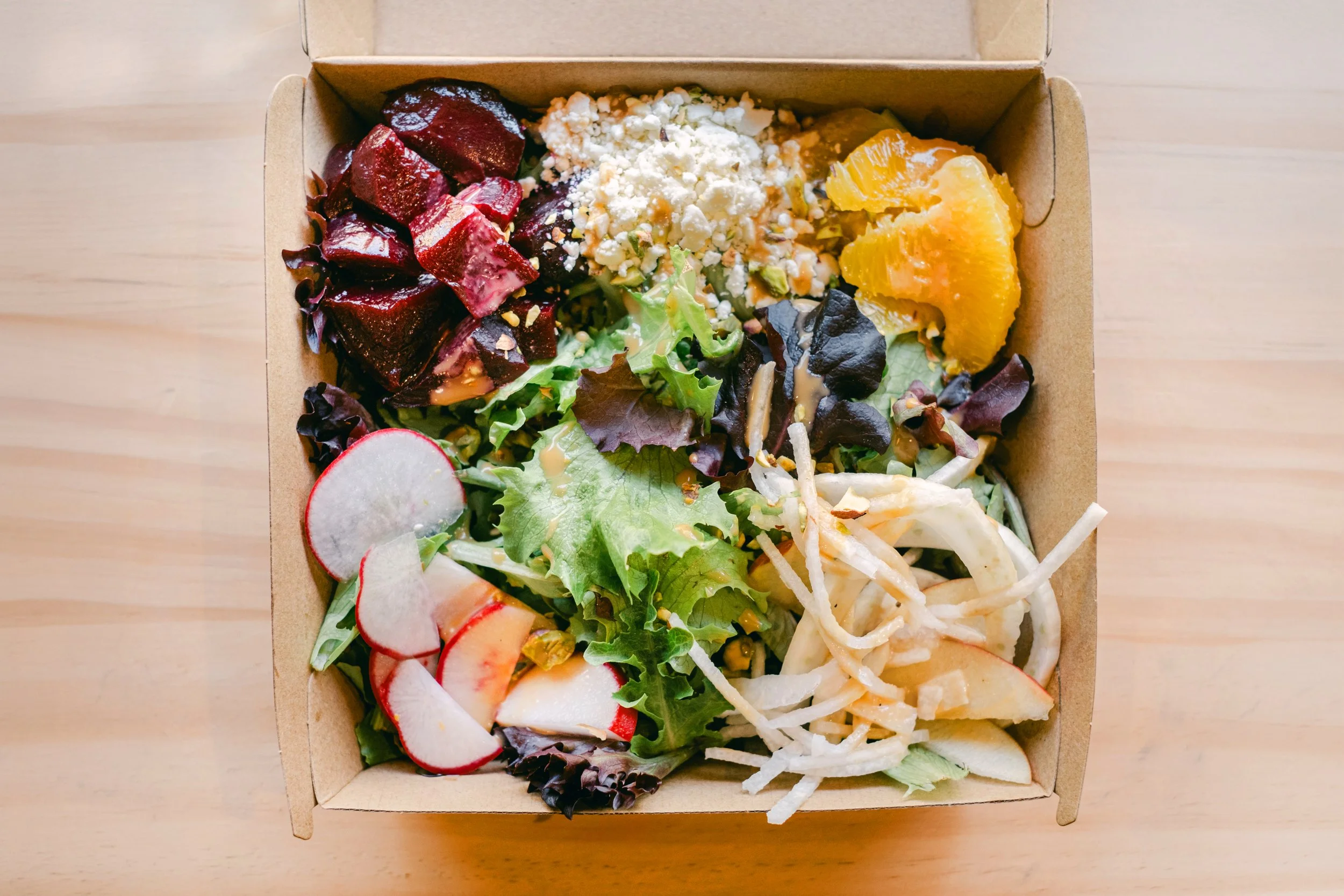 A cardboard takeout box filled with a salad containing mixed greens, sliced radishes, orange segments, shredded cheese, crumbled cheese, roasted beets, and shredded chicken.