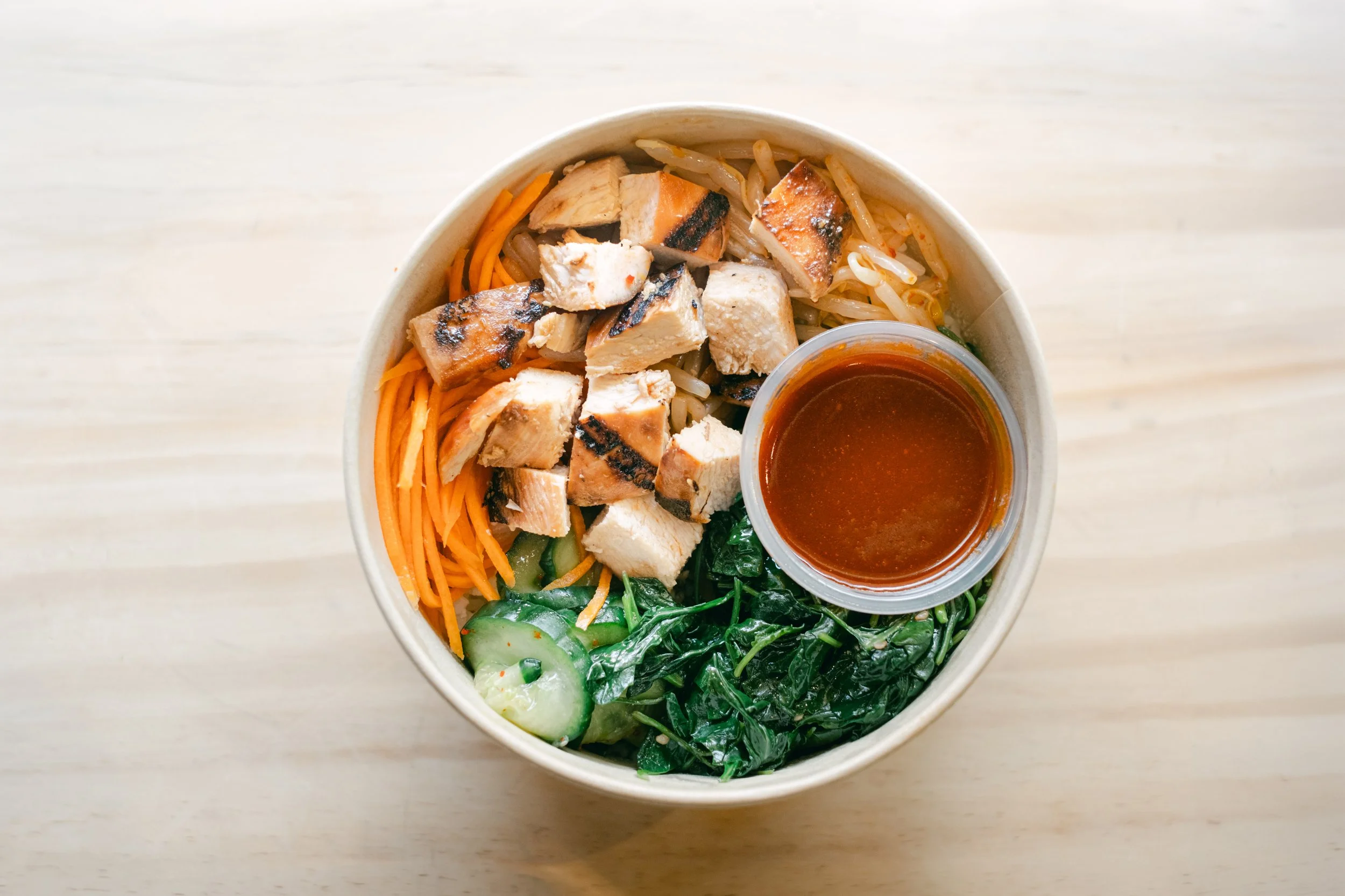 A bowl of Asian noodle salad topped with grilled chicken pieces and served with a side of spicy sauce.