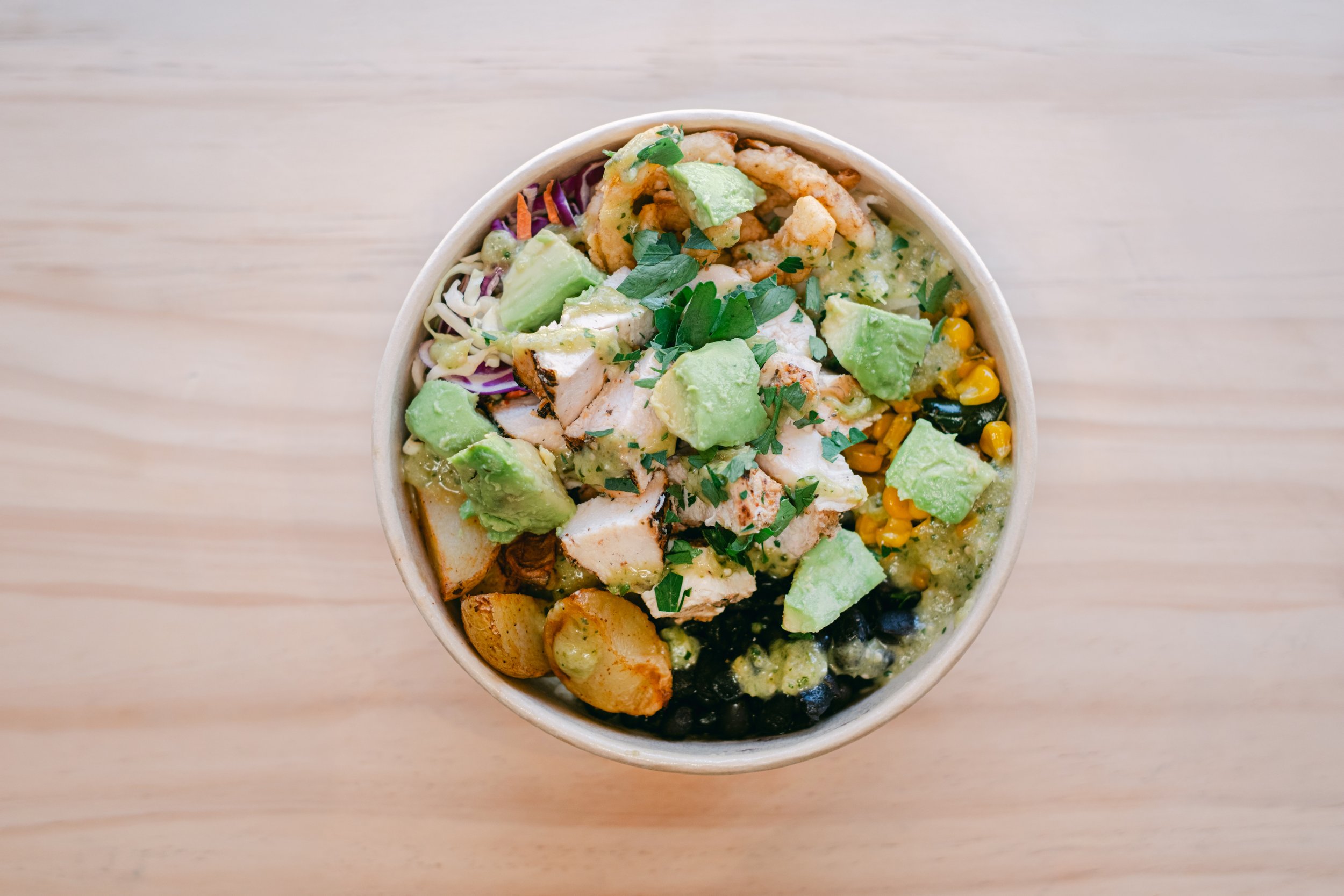 A bowl of colorful, topped with chopped herbs, avocado chunks, grilled chicken pieces, corn, and shredded vegetables on a light wooden surface.