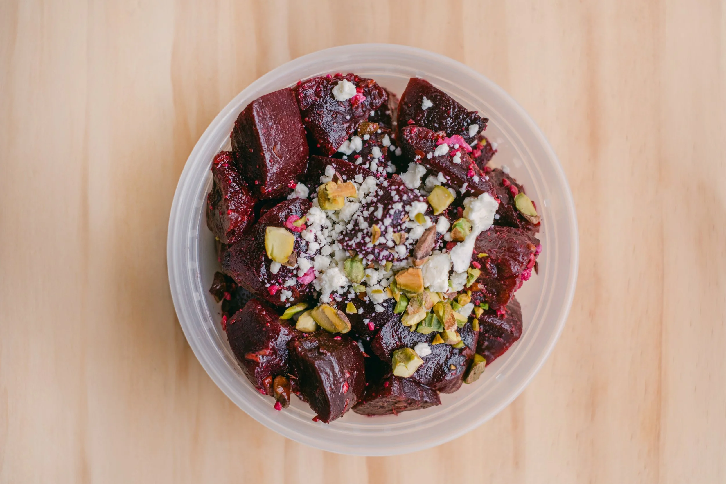 Close-up of a plastic container filled with a dessert made of dark red beetroot chunks garnished with chopped pistachios, crumbled white cheese, and pink and white sugar sprinkles, placed on a wooden surface.