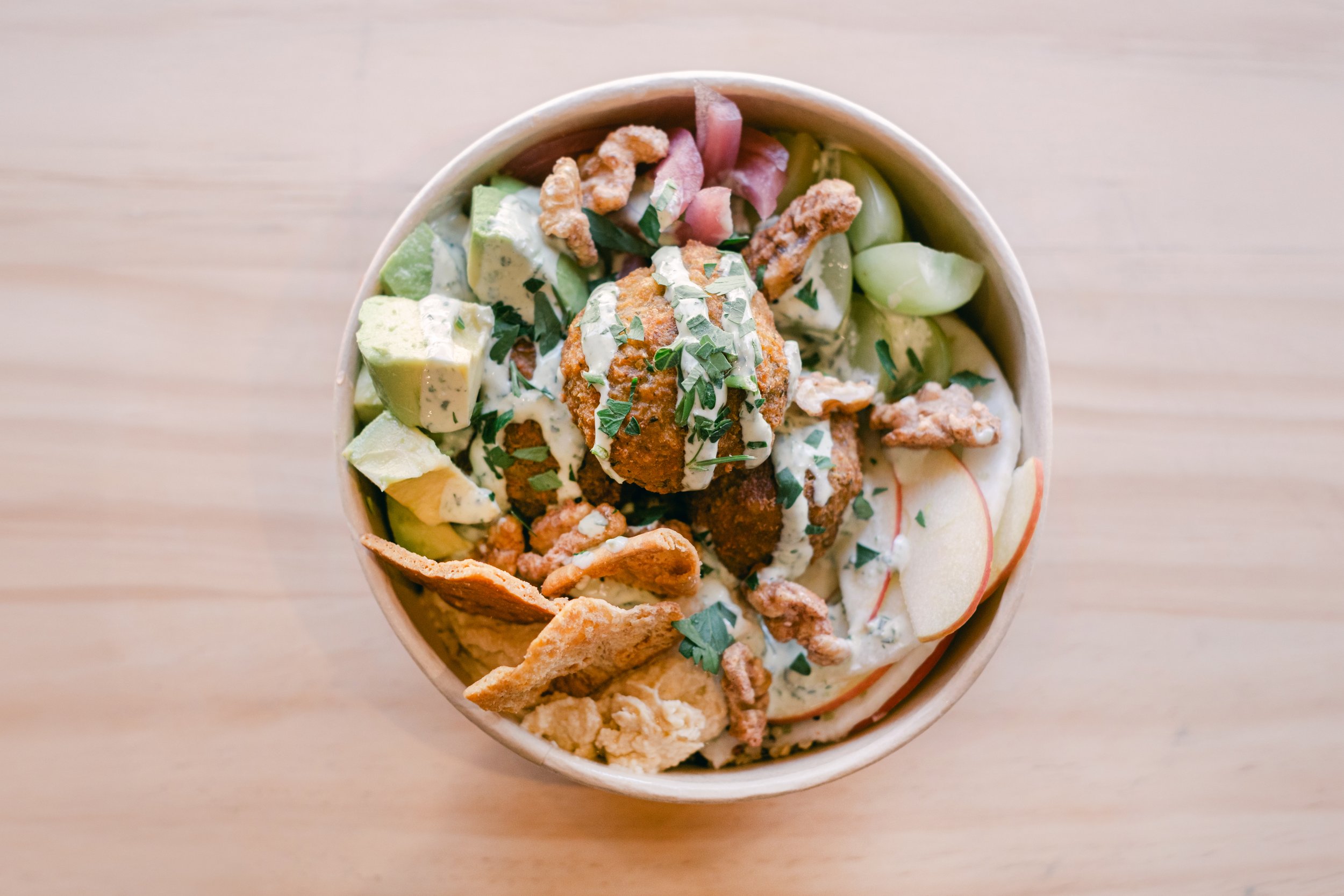 A bowl of salads with avocados, cherry tomatoes, chopped herbs, fried chicken pieces, and crackers on a light wood surface.