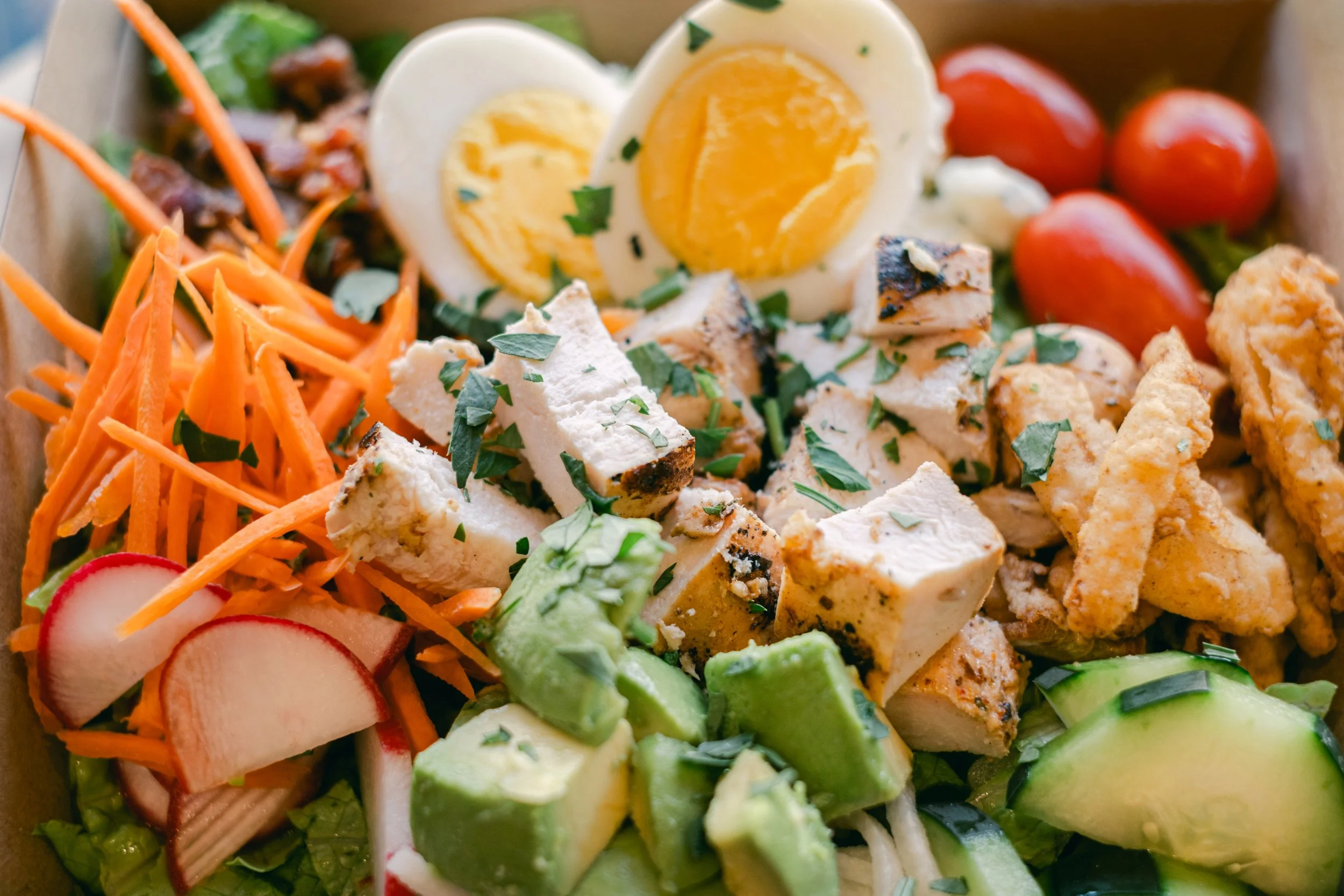 A close-up of a fresh salad with ingredients including sliced radishes, shredded carrots, cherry tomatoes, sliced grilled chicken, chopped hard-boiled eggs, sliced cucumbers, avocados, and leafy greens, garnished with chopped herbs.