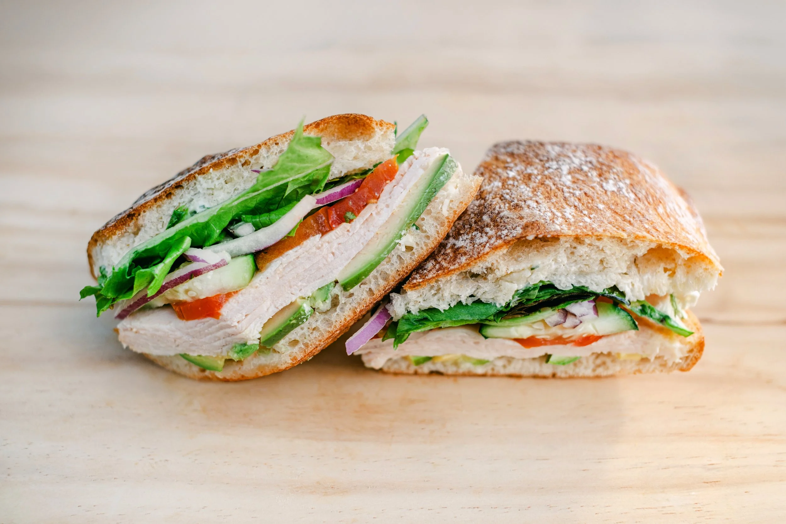 A close-up of a sliced turkey club sandwich with lettuce, tomato, cucumber, and onions on a toasted sub roll, on a light wooden surface.