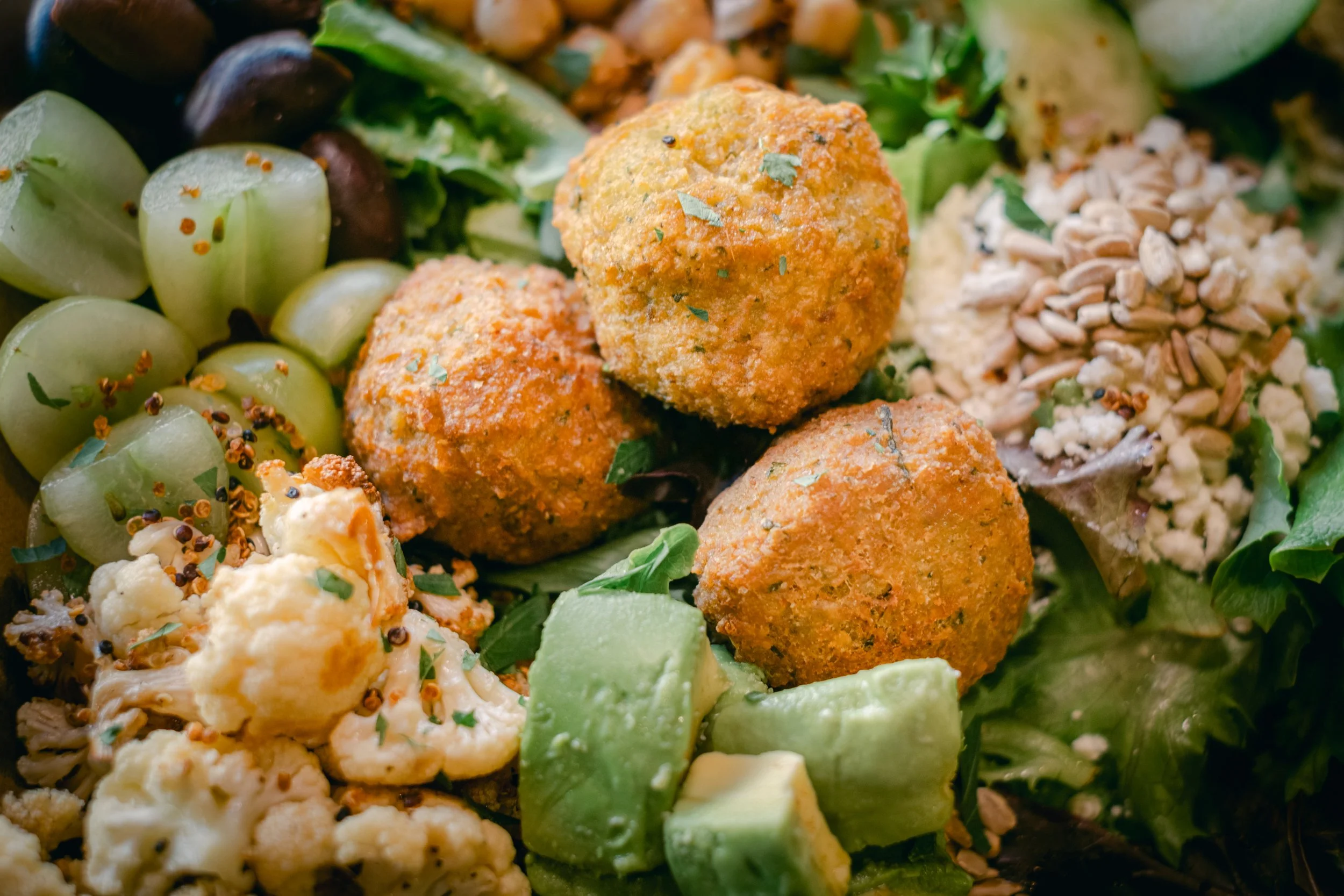 A close-up of a salad featuring falafel balls, chopped cucumbers, cauliflower, sunflower seeds, and mixed greens, garnished with herbs.