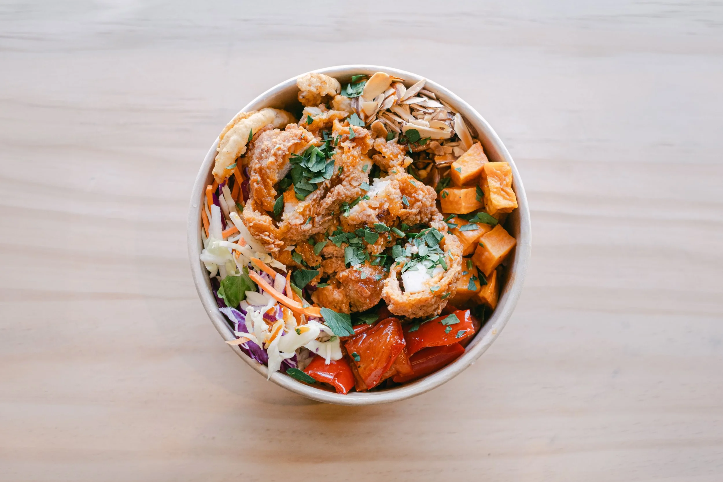 Overhead view of a bowl filled with chopped vegetables and fried chicken, garnished with chopped herbs, on a light wooden surface.