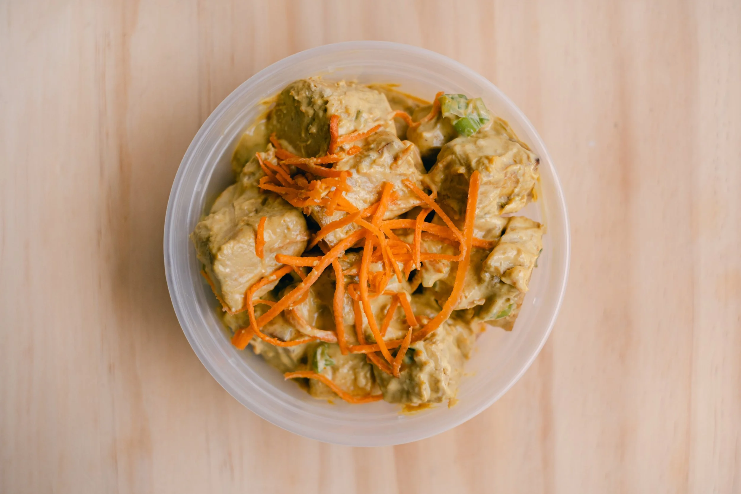 A clear plastic container filled with creamy chicken salad topped with shredded carrots and chopped green onions, placed on a light wooden surface.