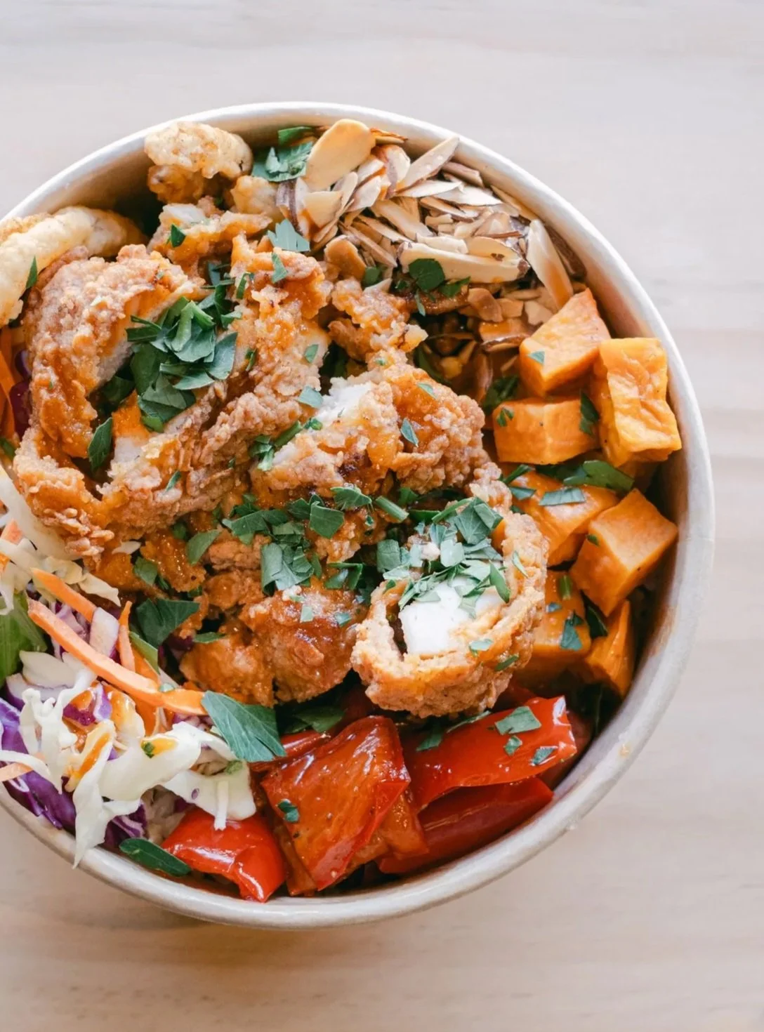 A bowl of Asian-inspired fried chicken with sweet potatoes, roasted red peppers, shredded cabbage, and chopped herbs.