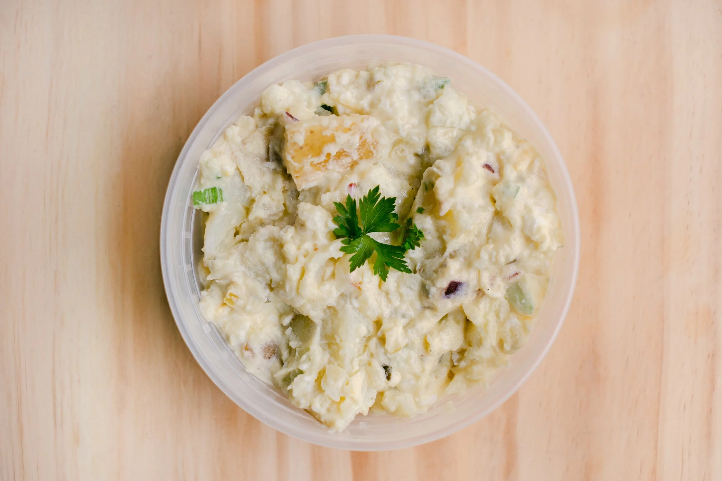 A plastic container filled with potato salad garnished with a sprig of parsley on a wooden surface.