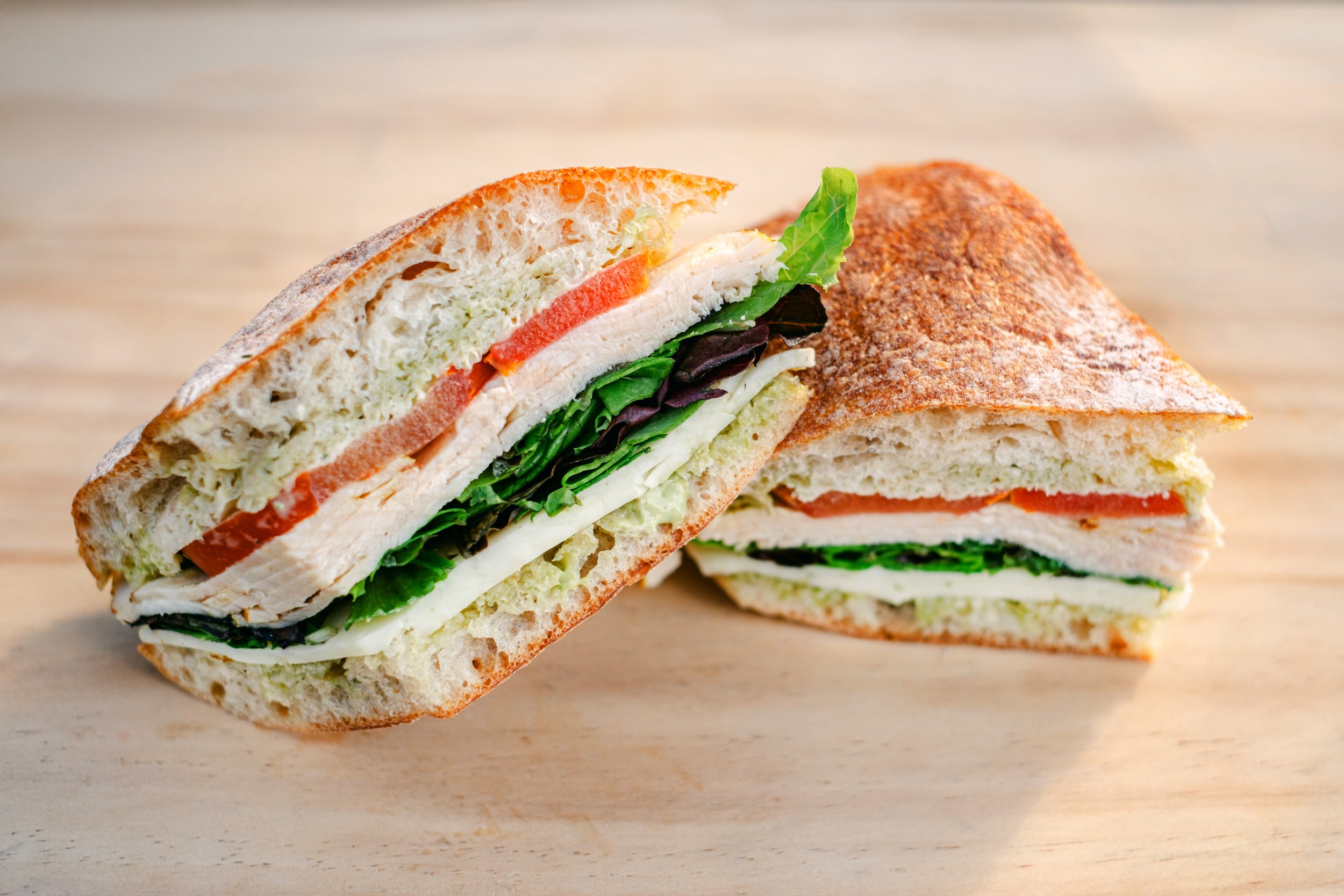 A close-up of a sandwich cut in half, showing lettuce, tomato, chicken, and sandwich bread on a light wooden surface.