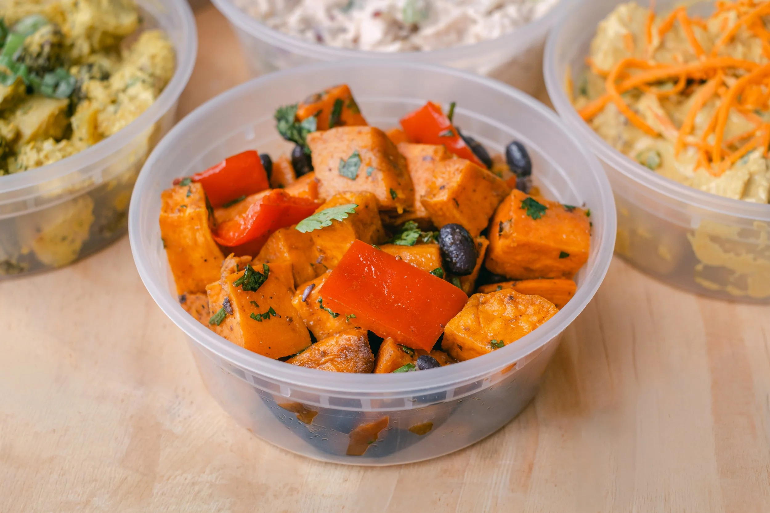 Close-up of three clear plastic containers filled with different types of salads or side dishes on a light wooden surface.