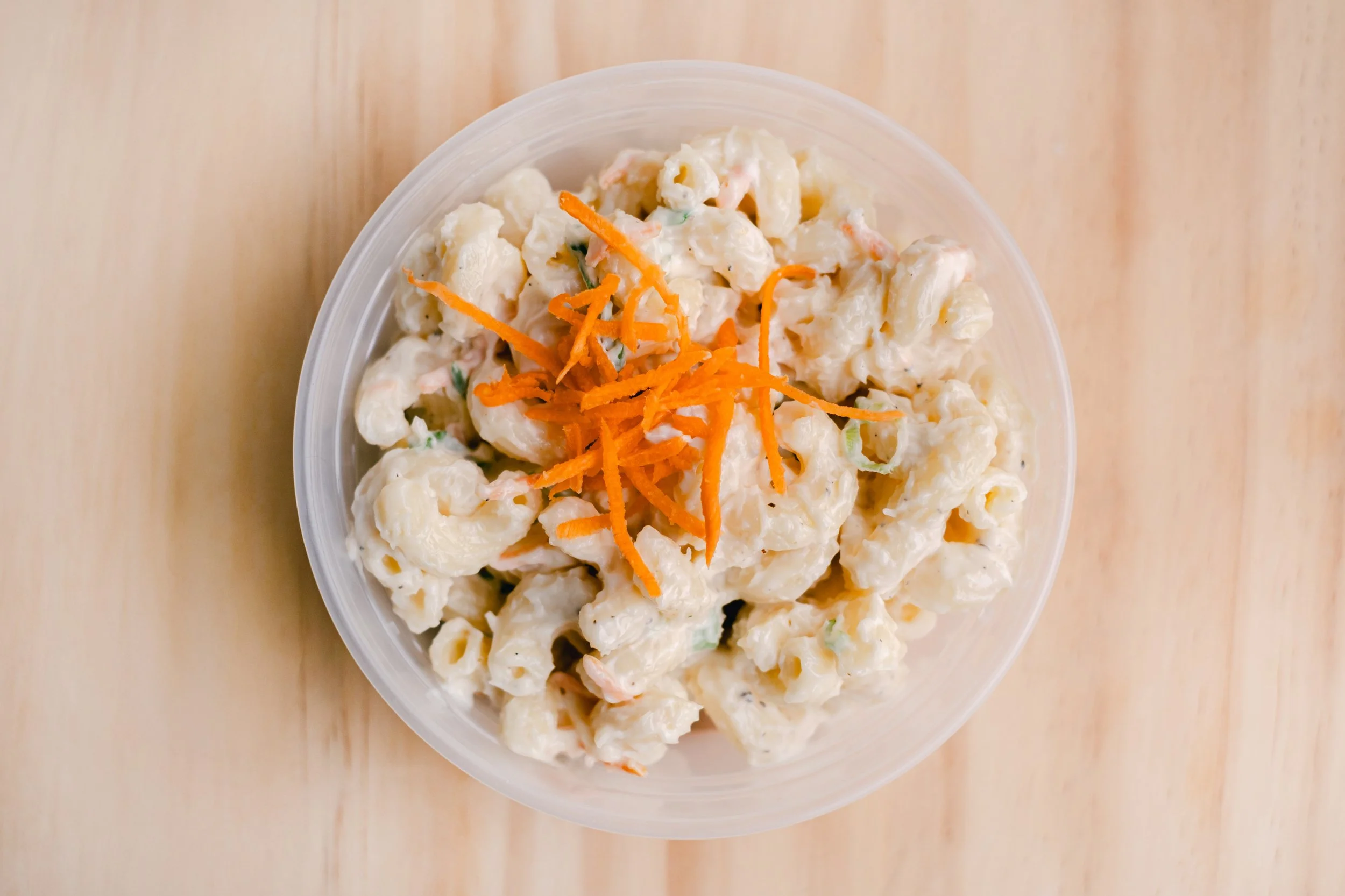 A clear plastic bowl filled with creamy potato salad topped with shredded carrots, placed on a wooden surface.