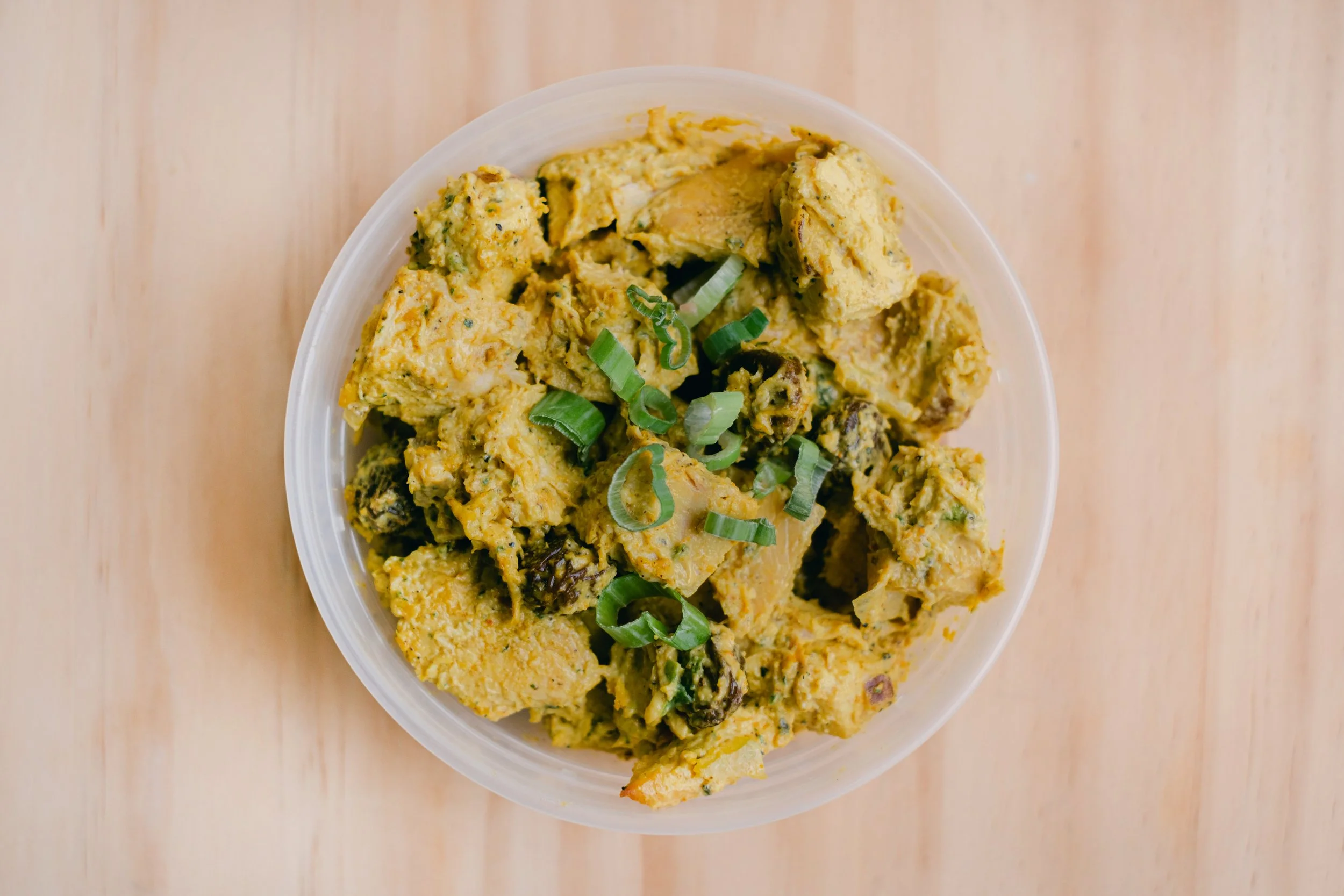 A close-up of a bowl filled with yellow curry containing chunks of chicken and vegetables, garnished with sliced green onions, placed on a wooden surface.
