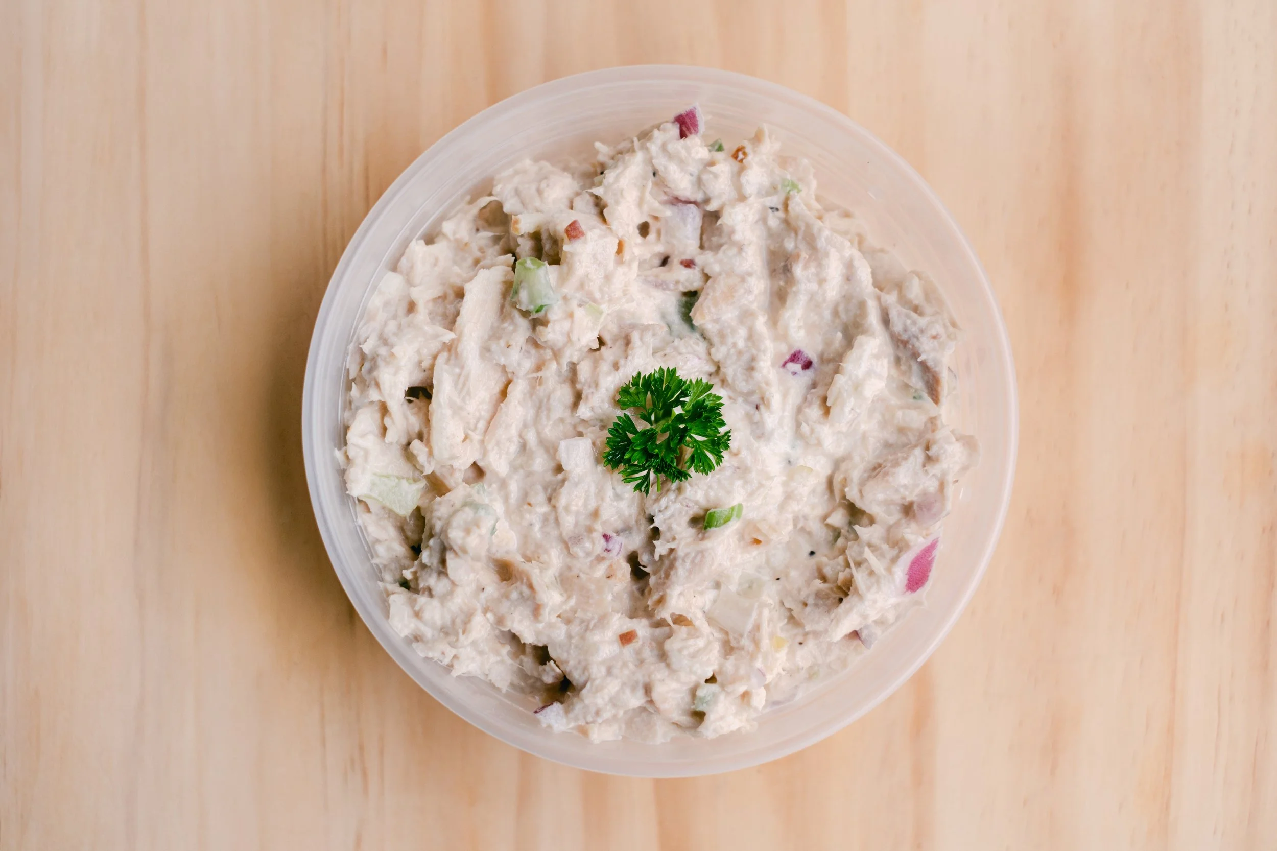 Close-up of a bowl filled with chicken salad garnished with a sprig of parsley, placed on a light wooden surface.