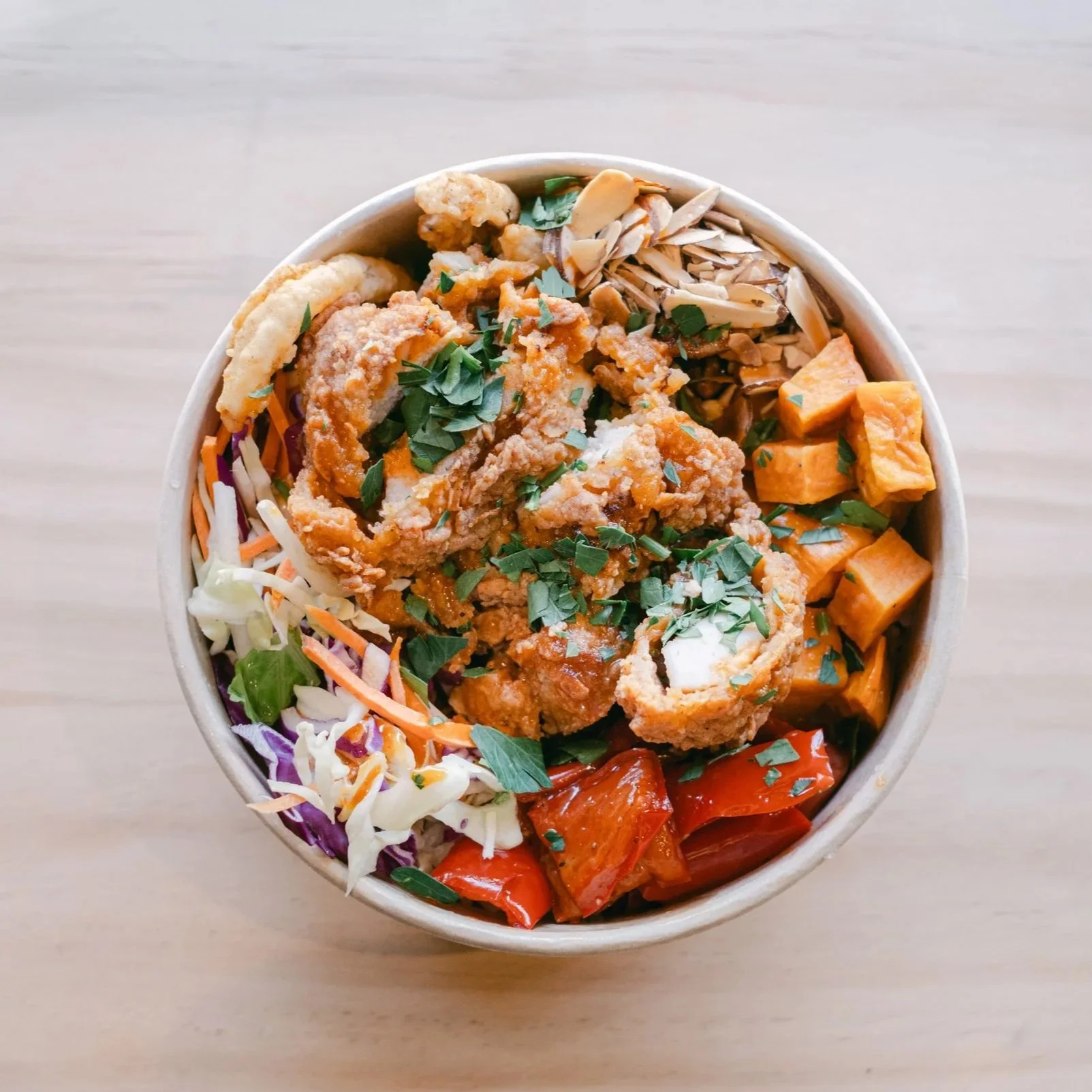 A bowl of Asian-inspired food with fried chicken, chopped herbs, shredded vegetables, tofu, and sliced mushrooms, served on a light wooden surface.