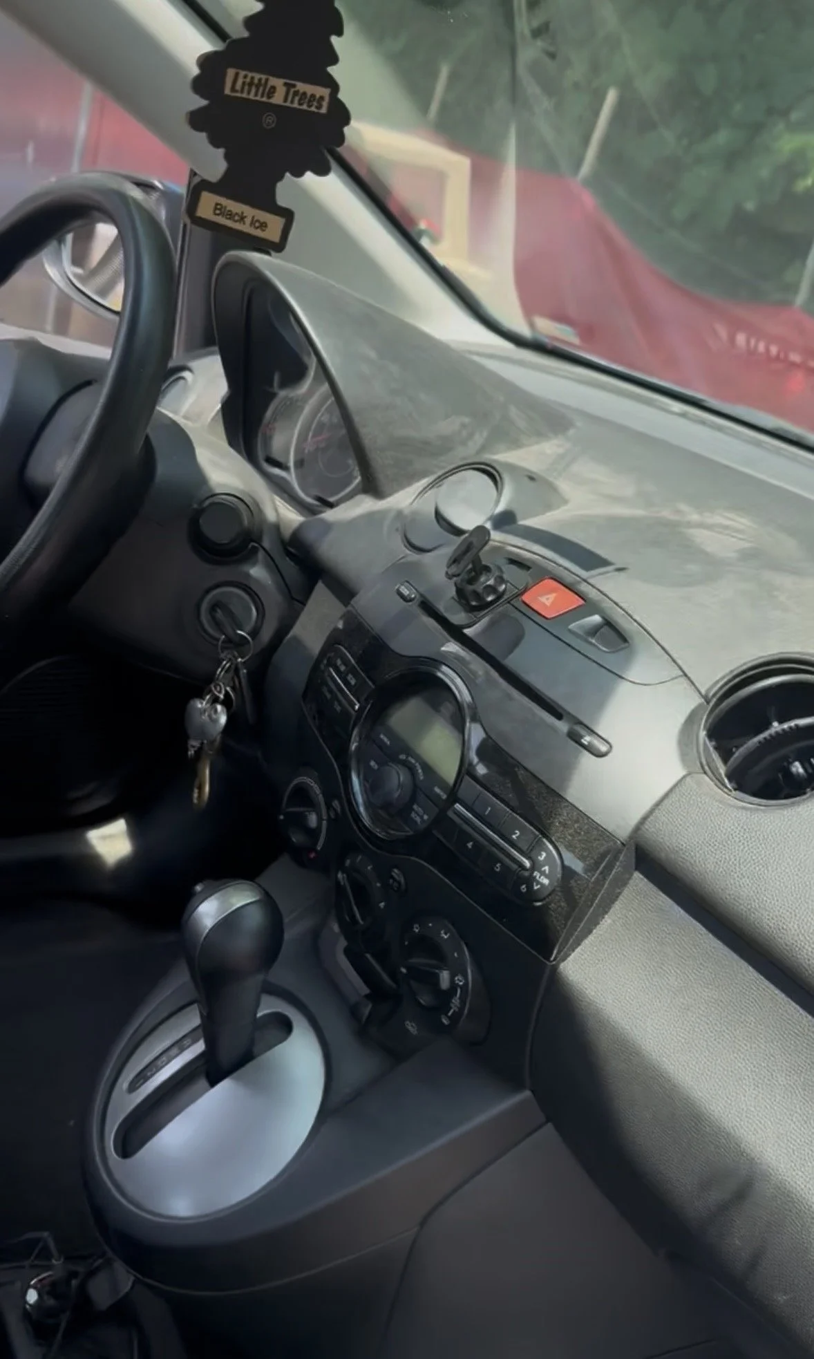 The interior of a car dashboard with a steering wheel, gear shift, and audio controls, and an air freshener hanging from the rearview mirror labeled 'Little Trees Black Ice.'
