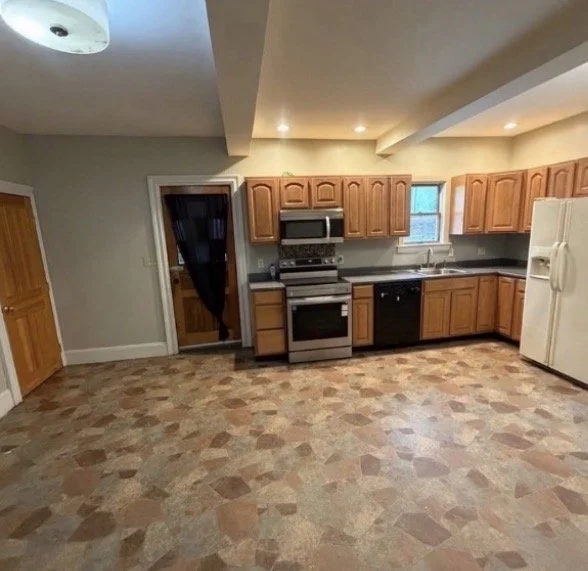 Empty kitchen with wooden cabinets, stainless steel stove and microwave, white refrigerator, and patterned tile floor.